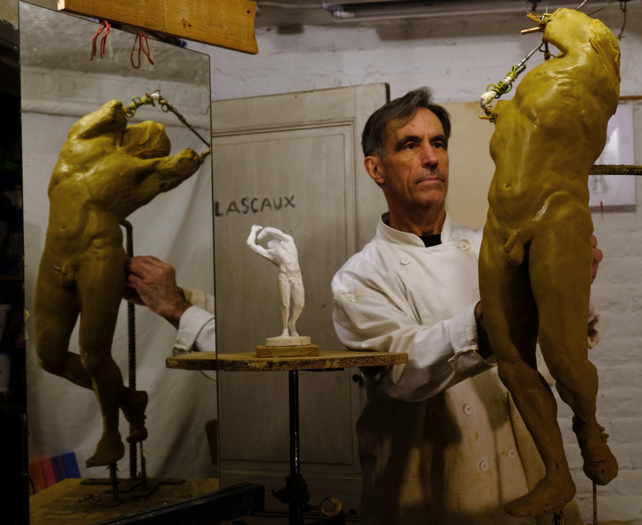 Un sculpteur Bruxellois en blouse blanche pose entre deux grandes figures masculin dorées dans son atelier, avec un modèle blanc en plâtre sur un piédestal et des outils de sculpture visibles en arrière-plan.