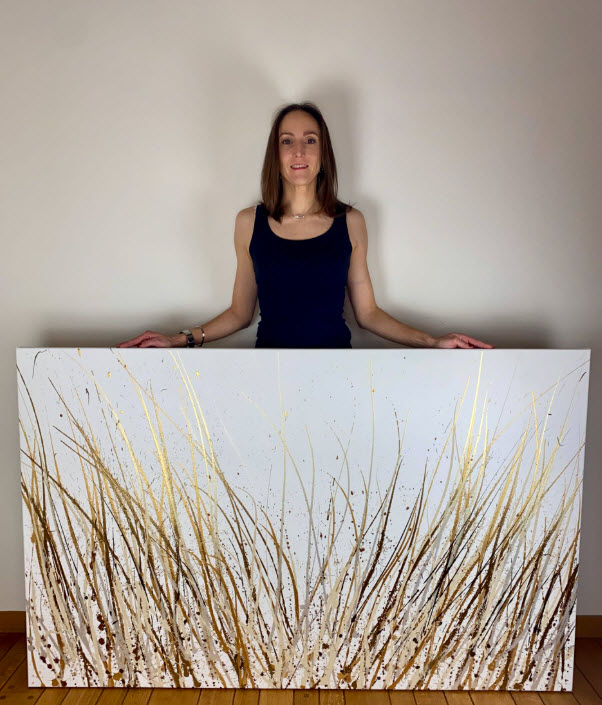 Lau Vago pose derrière un tableau abstrait aux traits de peinture dorée et brun sur fond blanc, dans un intérieur bruxellois.