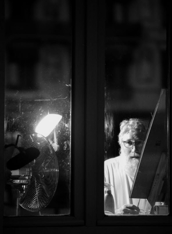 Portrait en noir et blanc de Boris Mestchersky, peintre, portant des lunettes et vêtu d'un t-shirt blanc, photographié à travers une vitre dans son atelier à Huy, avec un ventilateur et une lumière en arrière-plan.

Boris Mestchersky, artiste peintre hutois, dans son atelier le soir