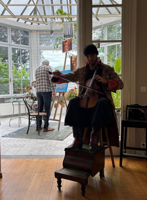 Un musicien joue du violoncelle assis sur un tabouret dans l'atelier Villa Sauvage, aux grandes baies vitrées, tandis qu'un peintre Boris Mestchersky travaille sur un chevalet en arrière-plan dans une ambiance de création partagée. Dialogue musical 2026.