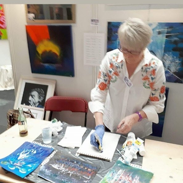 Nicole De Pauw, peintre, travaille à sa table d'atelier en portant des gants de protection, entourée de toiles colorées et de matériel de peinture. Artiste peintre Woluwe Saint Lambert