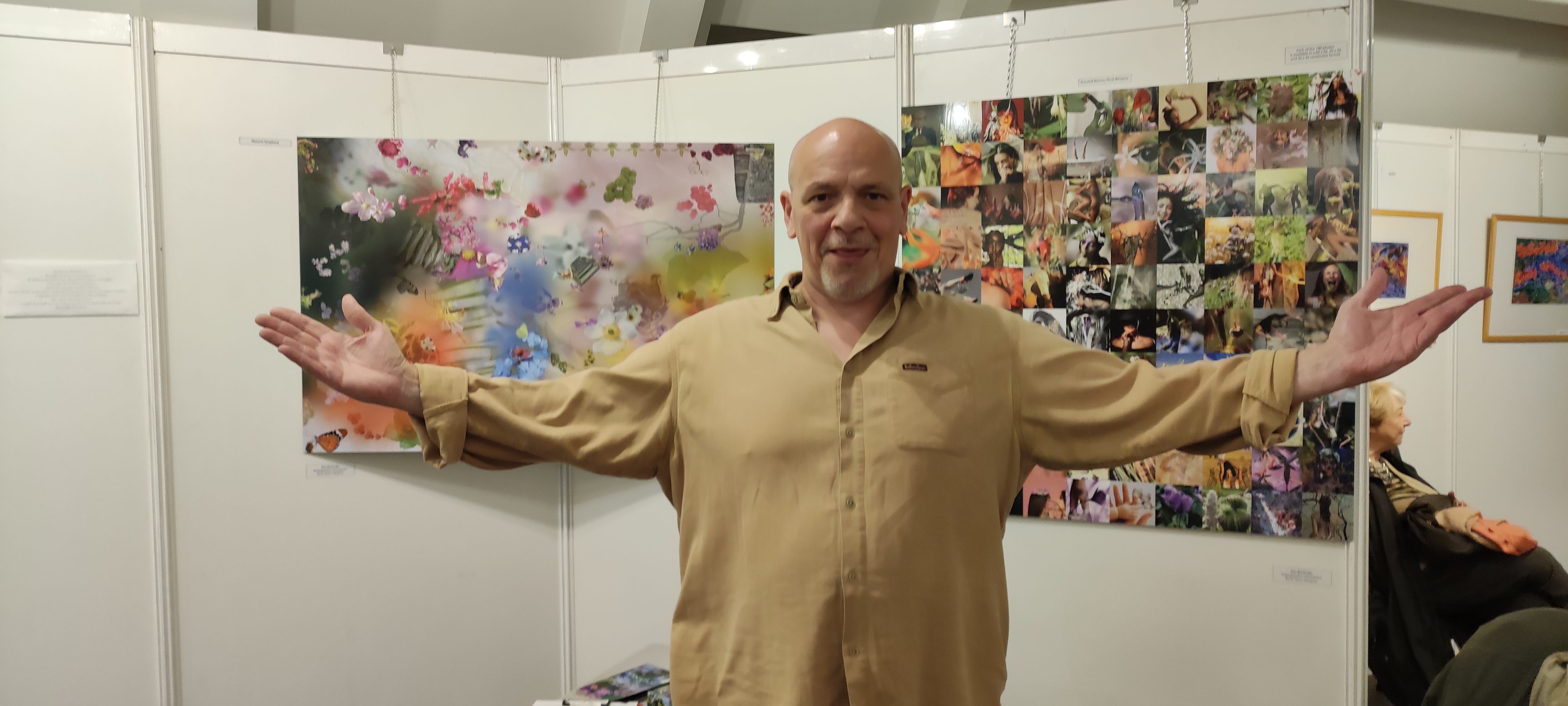 Un homme vêtu d'une chemise beige aux bras écartés pose devant un stand d'exposition présentant des photographies de fleurs et de portraits en couleur, dans un cadre de galerie ou de salon artistique.