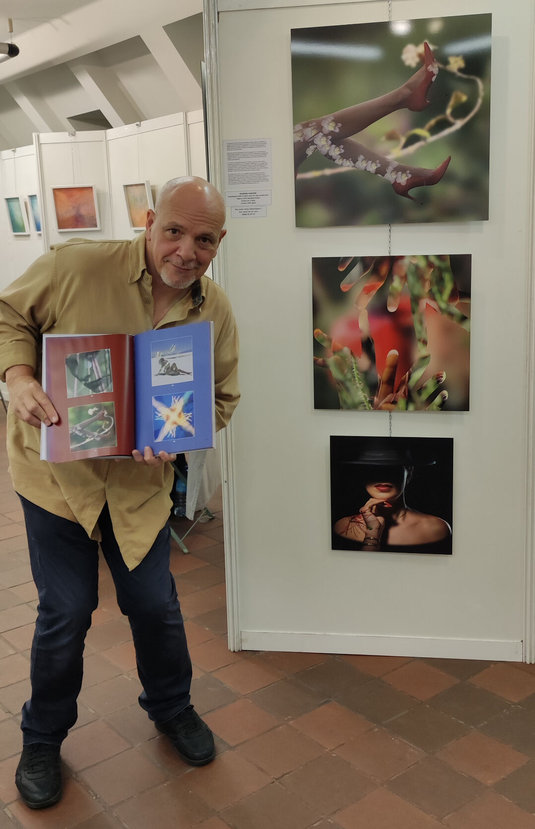 Un homme en chemise ocre tient un portfolio ouvert devant des photographies murales affichées dans une galerie bruxelloise, présentant des œuvres de nature et de portrait.