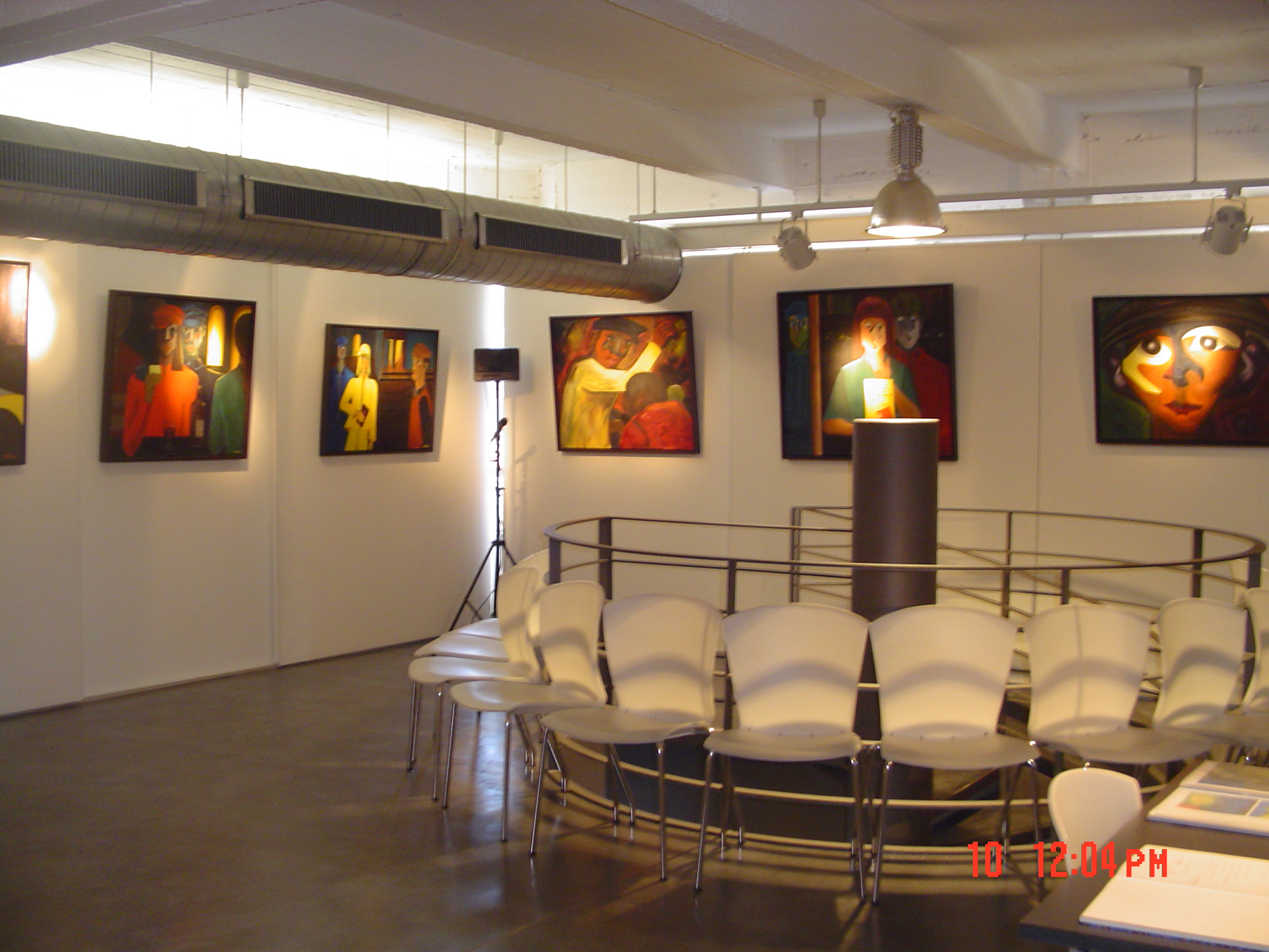 Intérieur de l'Espace Wallonie à Nivelles avec des tableaux expressionnistes aux tons chauds (rouges, oranges, jaunes) accrochés sur les murs blancs, des chaises blanches disposées face à une estrade circulaire, et une suspension métallique au plafond.
