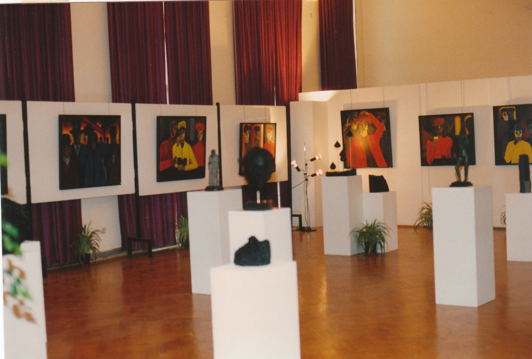 Vue d'ensemble d'une exposition bruxelloise présentant des peintures expressionnistes aux couleurs chaudes (rouge, orange, jaune) accrochées sur des panneaux blancs, avec des sculptures noires sur des socles blancs disposés au sol, sous des rideaux bordeaux et des éclairages ponctuels.