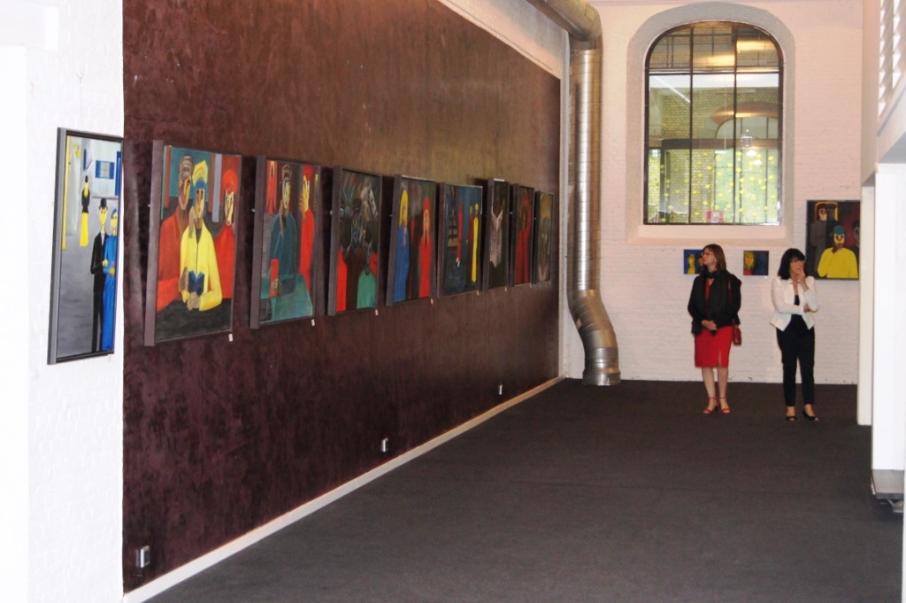 Intérieur d'une galerie bruxelloise avec mur bordeaux présentant une série de peintures de portraits aux couleurs vives (rouge, bleu, jaune), deux visiteuses en conversation au centre de l'espace, fenêtre arquée et tuyauterie métallique visibles.