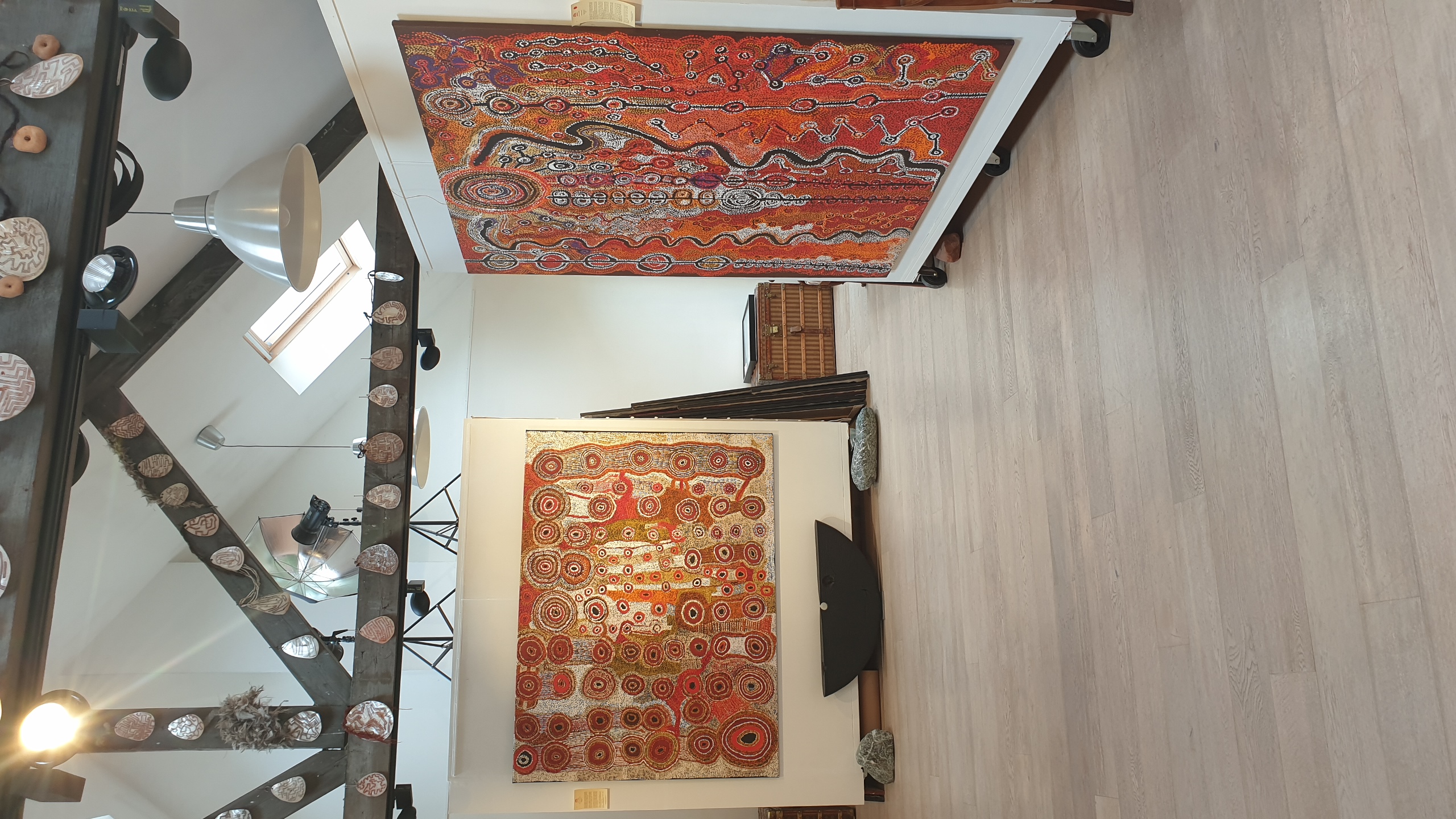 Galerie Aboriginal Signature à Bruxelles. Présentant deux grands textiles autochtones australiens aux motifs géométriques rouges et orangés accrochés aux murs, décorée de paniers tressés suspendus et d'éléments de mise en scène minimaliste.