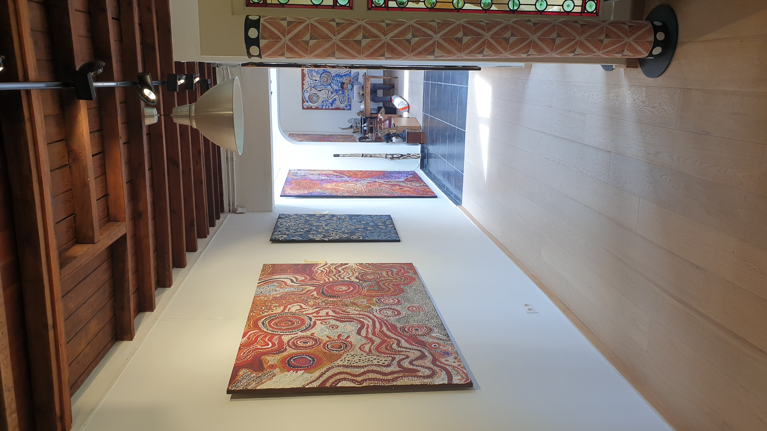 Salle d'exposition de la galerie bruxelloise Aboriginal Signature avec poutre apparente en bois, parquet clair, murs blancs accueillant des toiles abstraites aux motifs géométriques et patterns rouges, bleus et multicolores, sculptures décoratives sur les côtés. photographie: Jérôme Selosse. Visite en 2021.