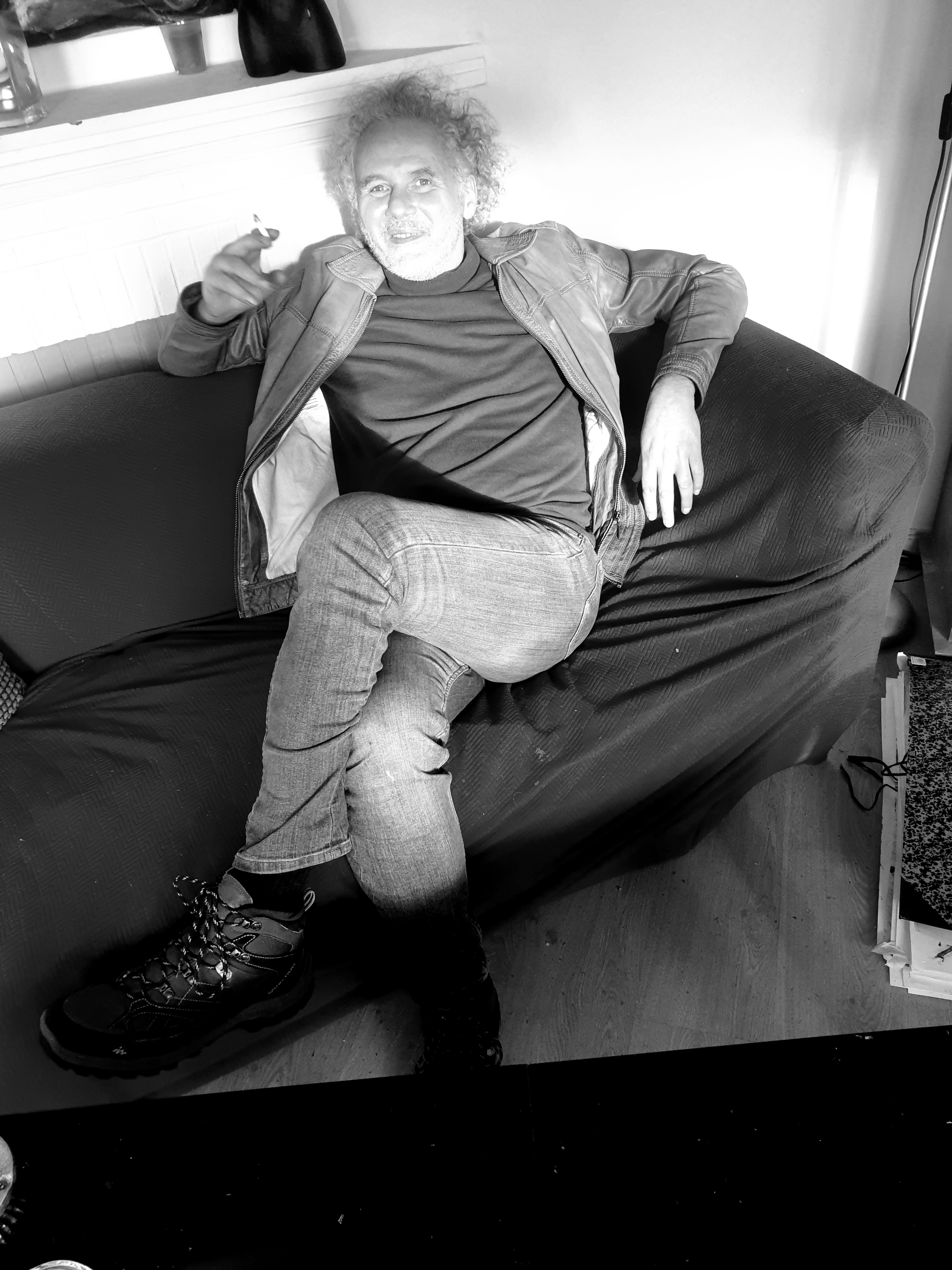 Portrait en noir et blanc de Jérôme Selosse, artiste basé à Bruxelles, assis sur un canapé, une jambe croisée, tenant une cigarette, regardant le spectateur avec un sourire.