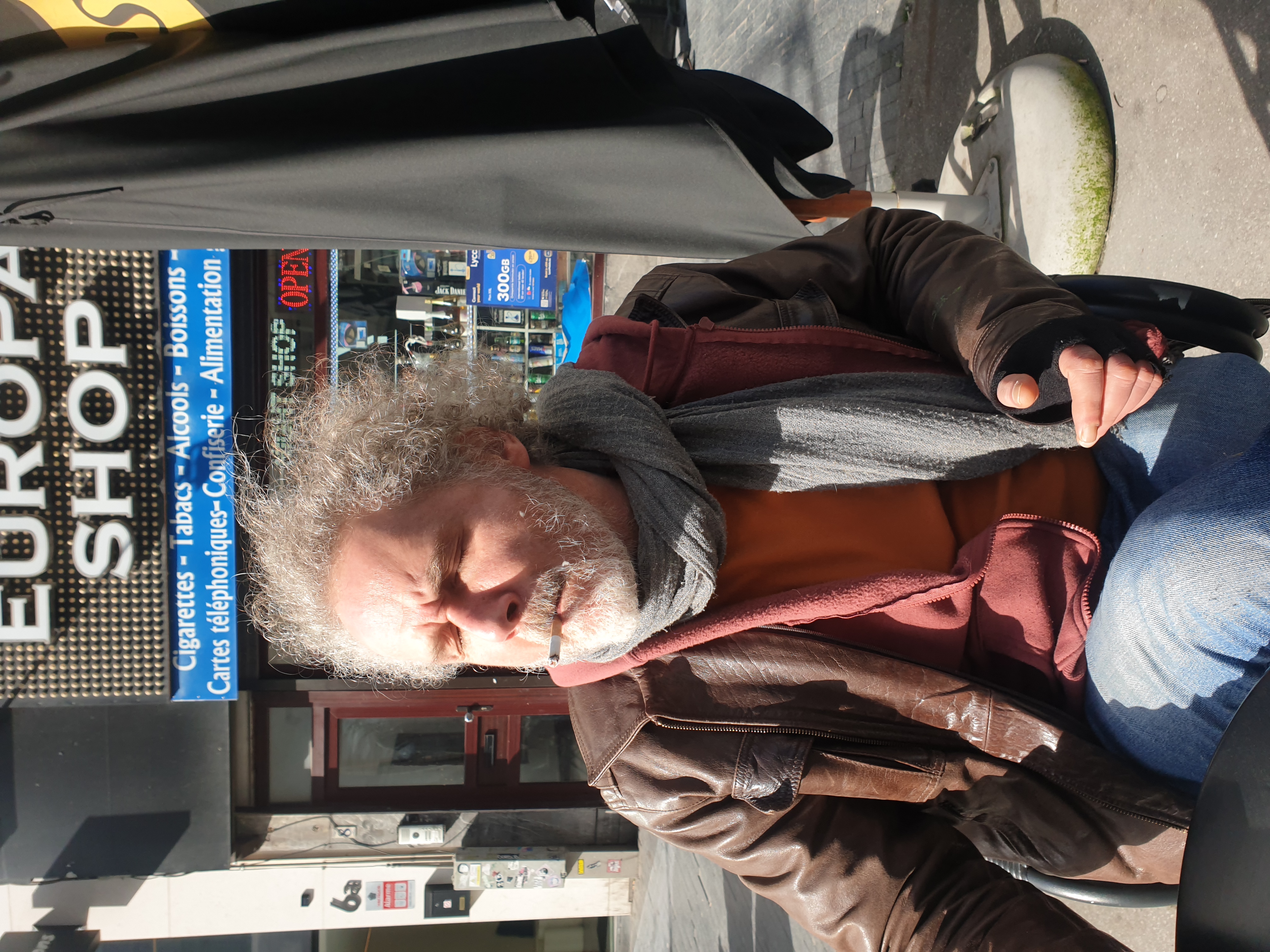 Portrait de Jérôme Selosse, artiste plasticien et illustrateur basé à Bruxelles, assis en rue devant un commerce, vêtu d'une veste marron et d'un foulard gris.
