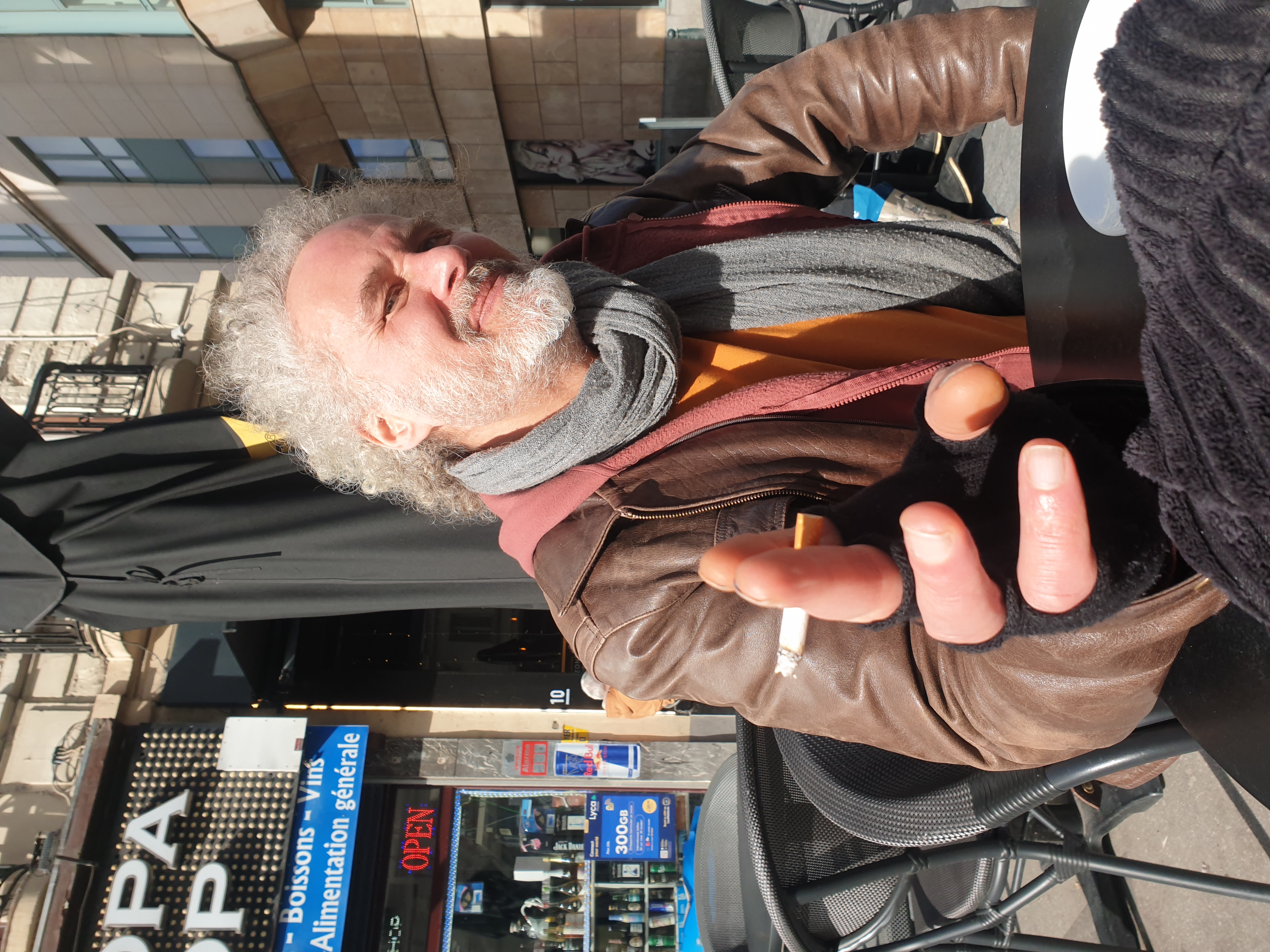 Portrait de Jérôme Selosse, artiste plasticien et illustrateur belge, assis en terrasse dans une rue bruxelloise, portant une veste marron en cuir et un foulard gris, gesticulant de manière expressive.