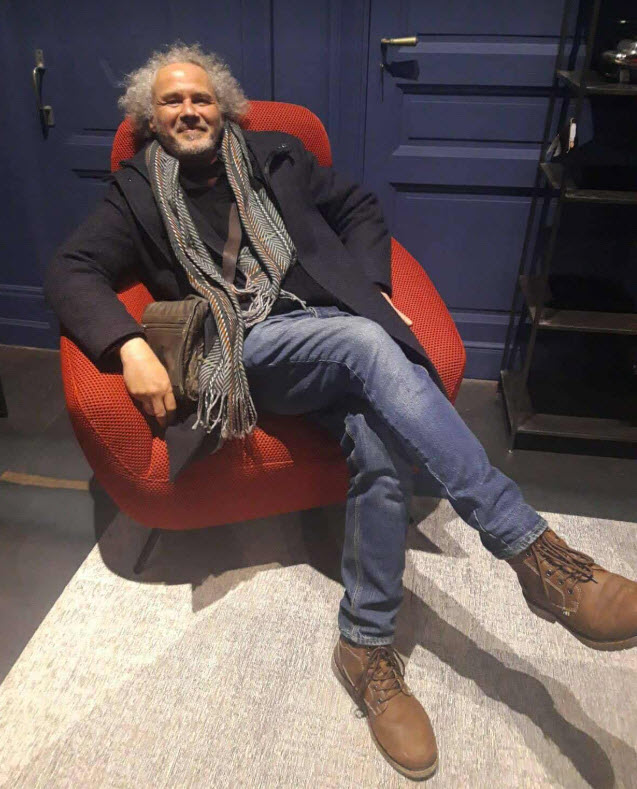 Portrait de Jérôme Selosse, artiste plasticien et illustrateur basé à Bruxelles, assis sur un fauteuil rouge vif dans un intérieur contemporain, vêtu d'un blazer noir et d'un foulard gris, souriant à la caméra.