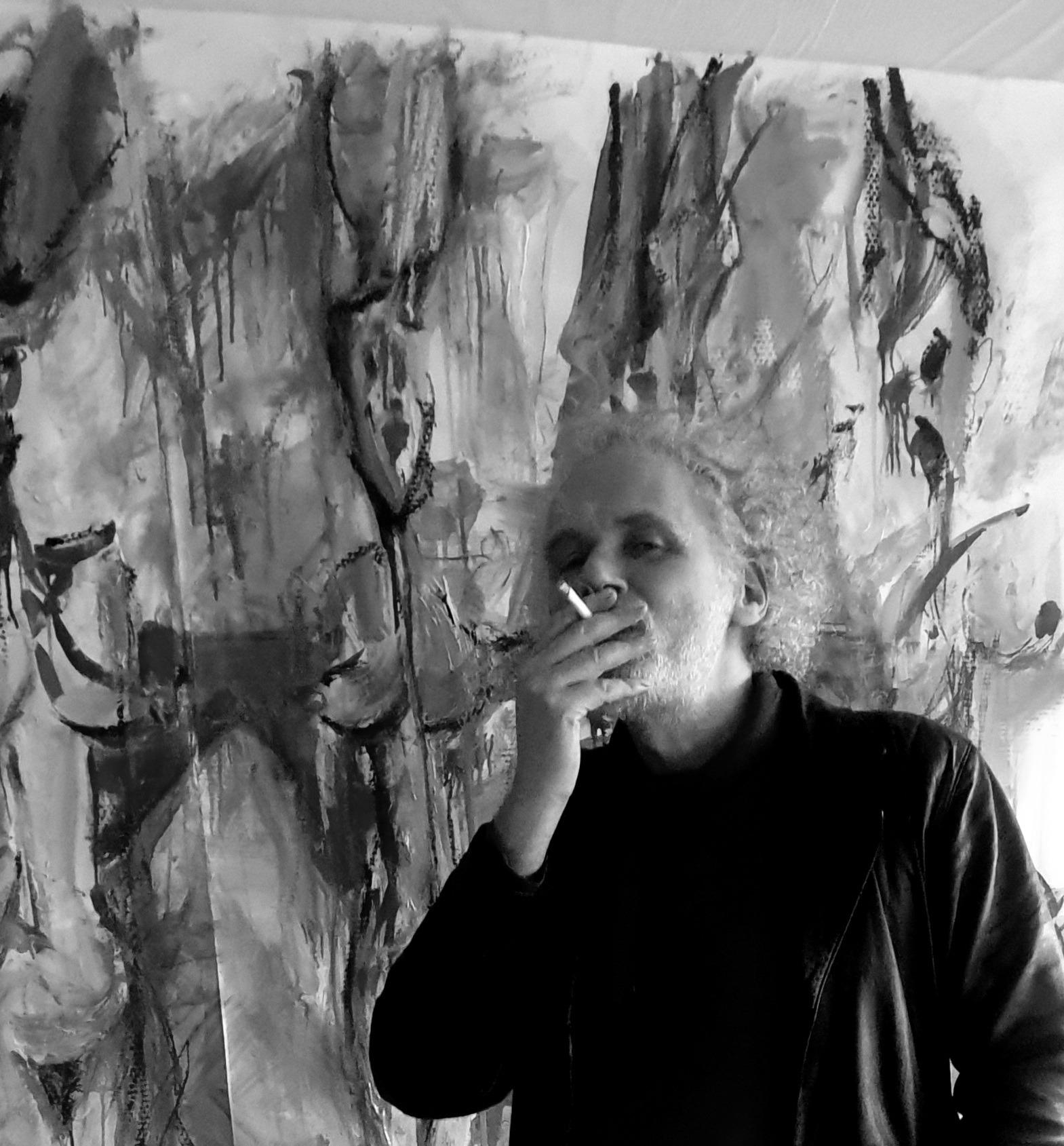 Portrait en noir et blanc de Jérôme Selosse, artiste plasticien bruxellois, posant devant une peinture abstraite caractérisée par des traits verticaux dynamiques et des textures gestuelles monochromes.