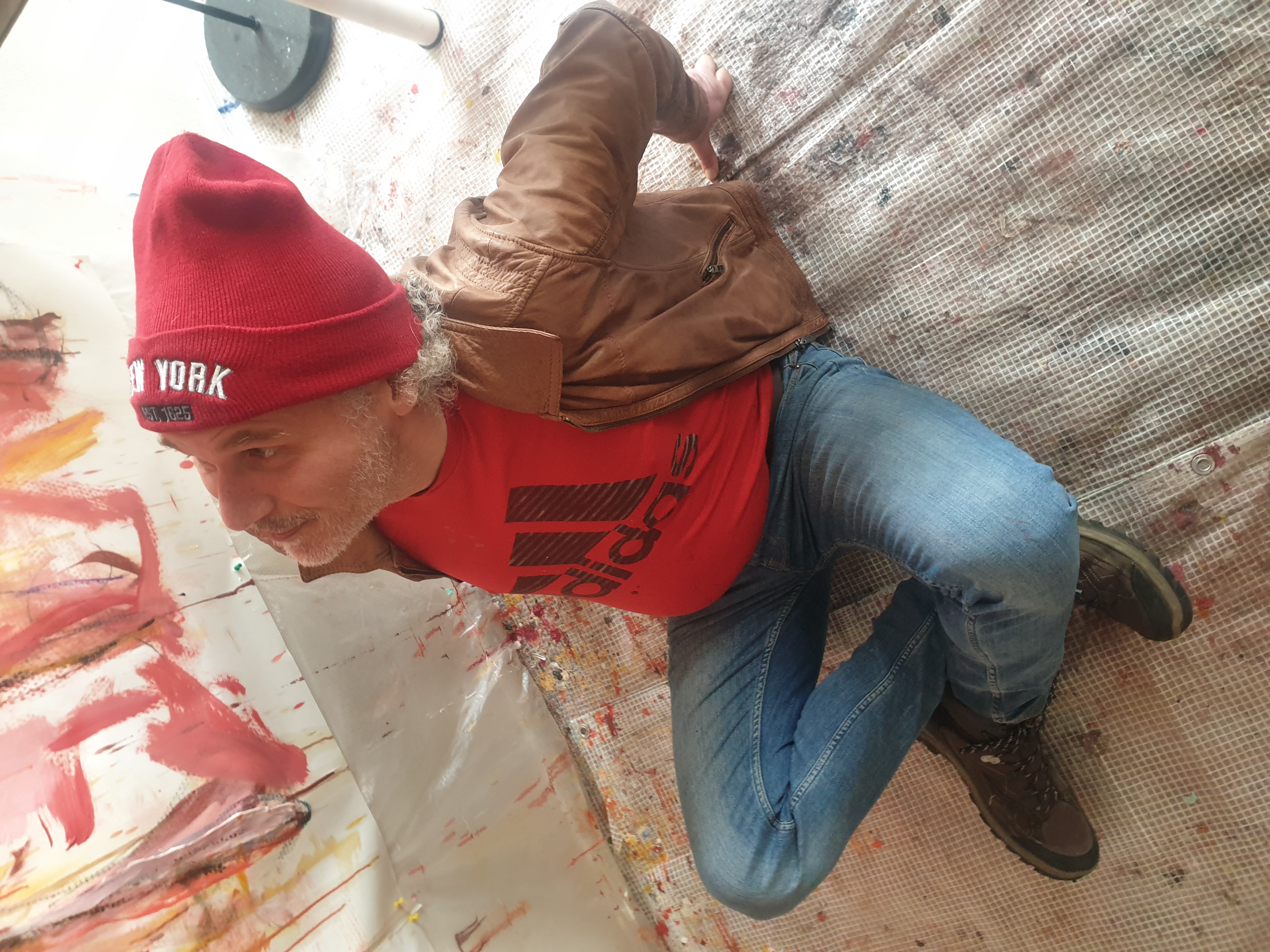 L'artiste plasticien Jérôme Selosse, portant un bonnet rouge et une veste marron, s'agenouille sur le sol recouvert de taches colorées face à une grande toile blanche aux traits rouges, orangés et jaunes, capturant son processus de création artistique.