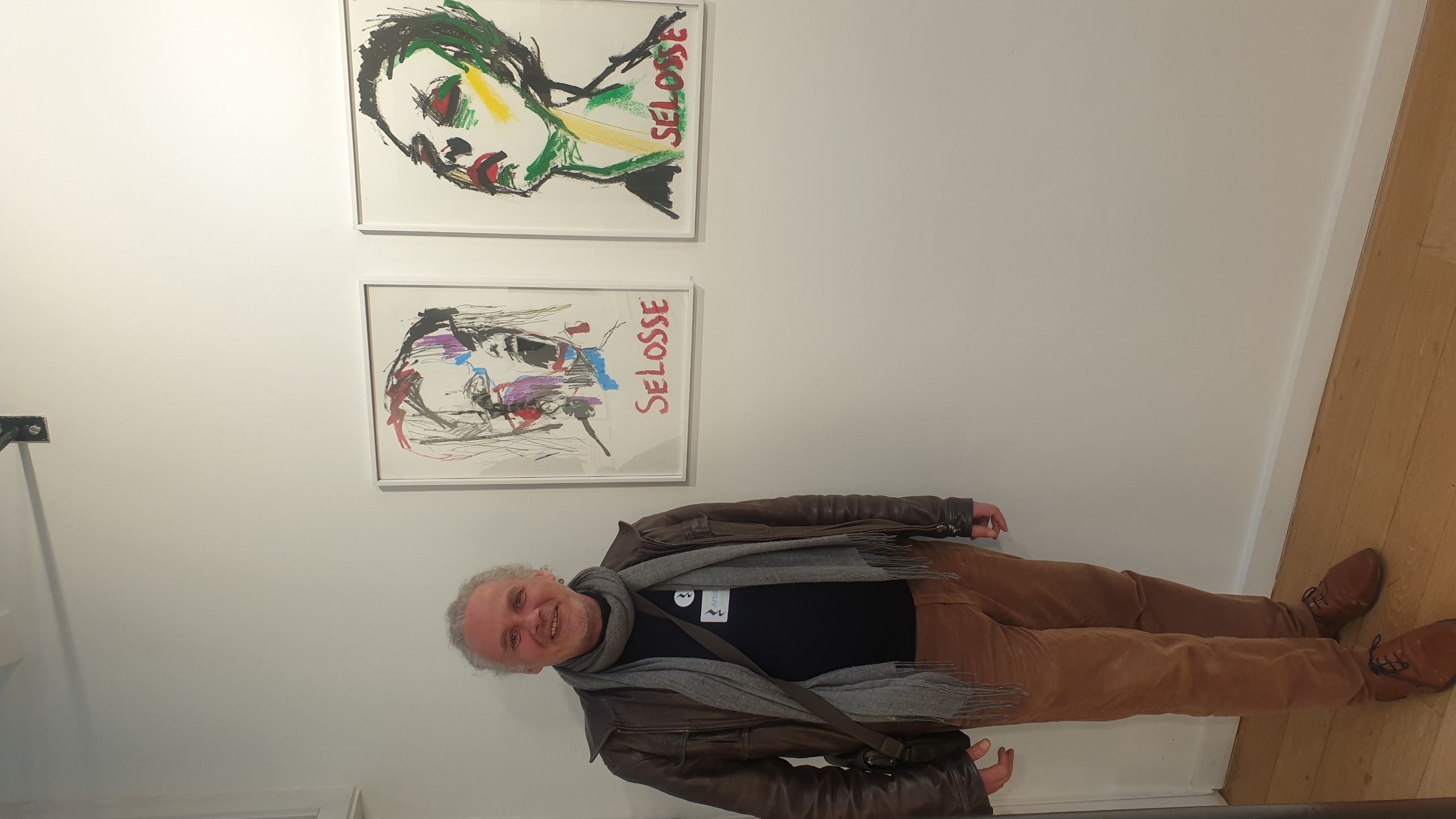 L'artiste Jérôme Selosse pose souriant face à deux dessins de portraits expressionnistes marqués du texte 'SELOSSE', exposés en galerie blanche. GALERIE Vertige Expo