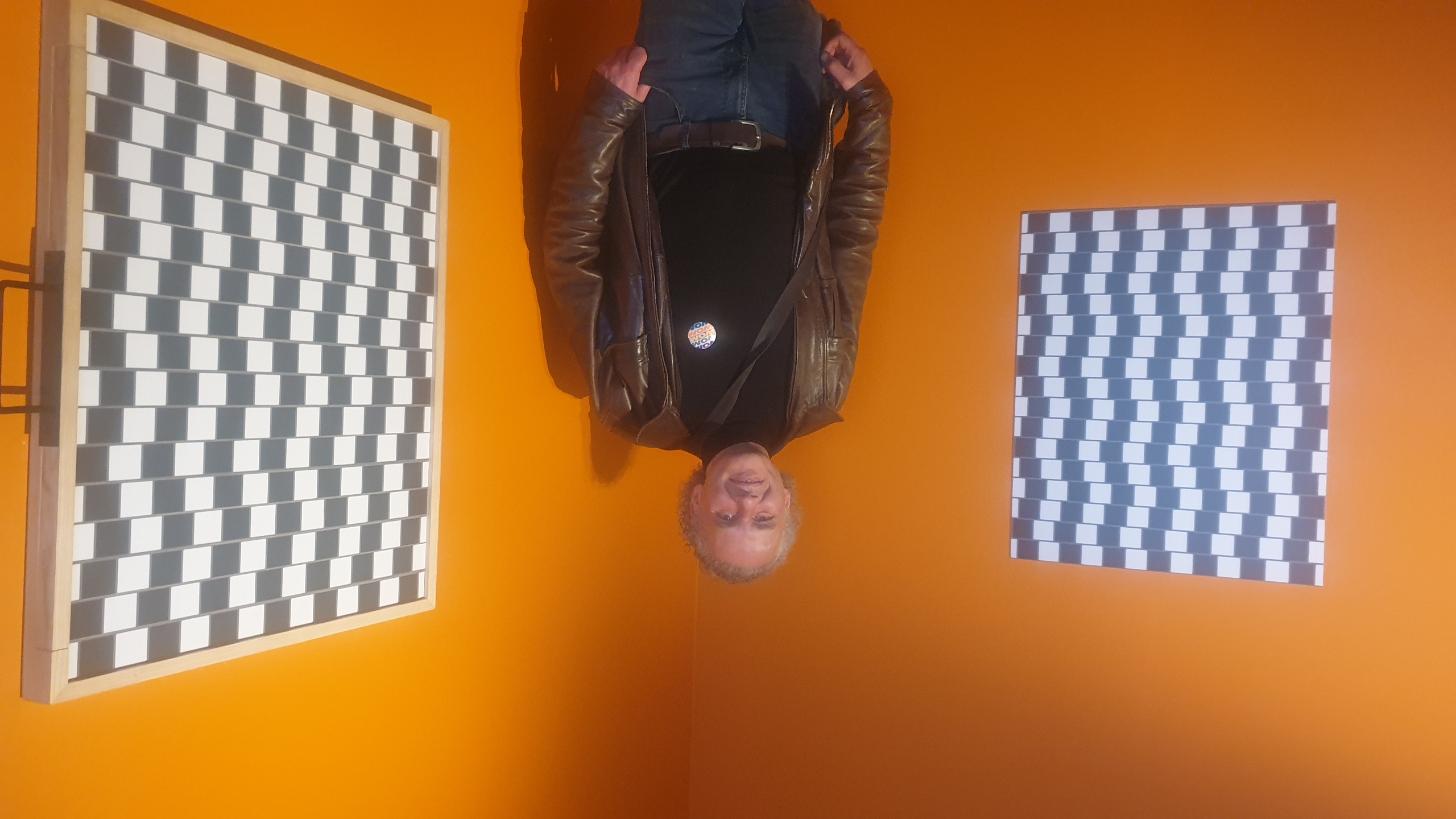 Jérôme Selosse en veste de cuir marron se tient debout au centre de la galerie des illusions au Wom. Des murs orange vif, encadré par deux grandes œuvres d'art abstrait aux motifs géométriques noir, blanc et gris. Wom de bruxelles.