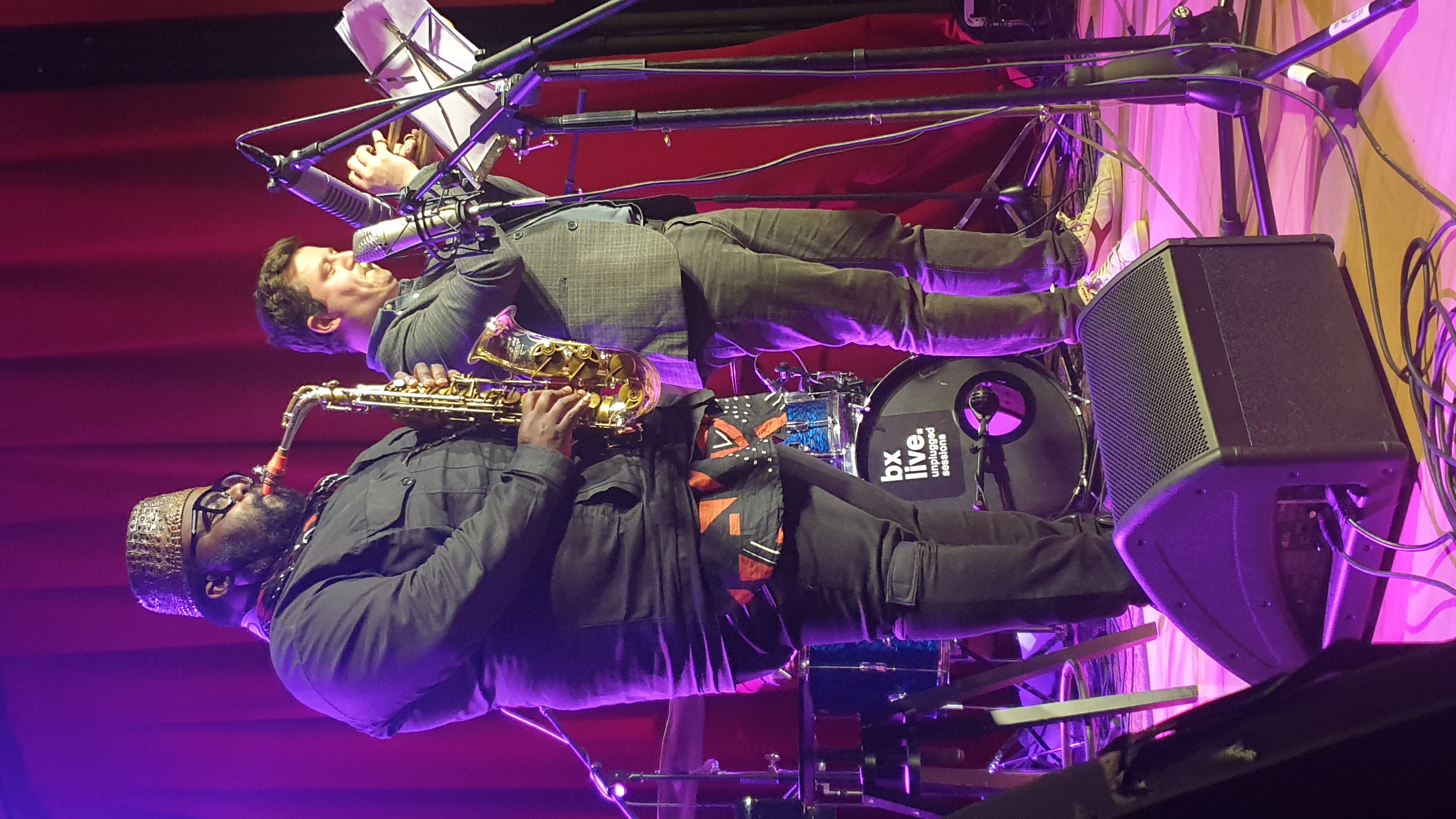 Deux musiciens bruxellois sur scène au Sounds Jazz club sous éclairage pourpre : un saxophoniste en chapeau doré jouant à gauche, un chanteur au micro à droite, devant un rideau rouge, lors d'une session acoustique.
