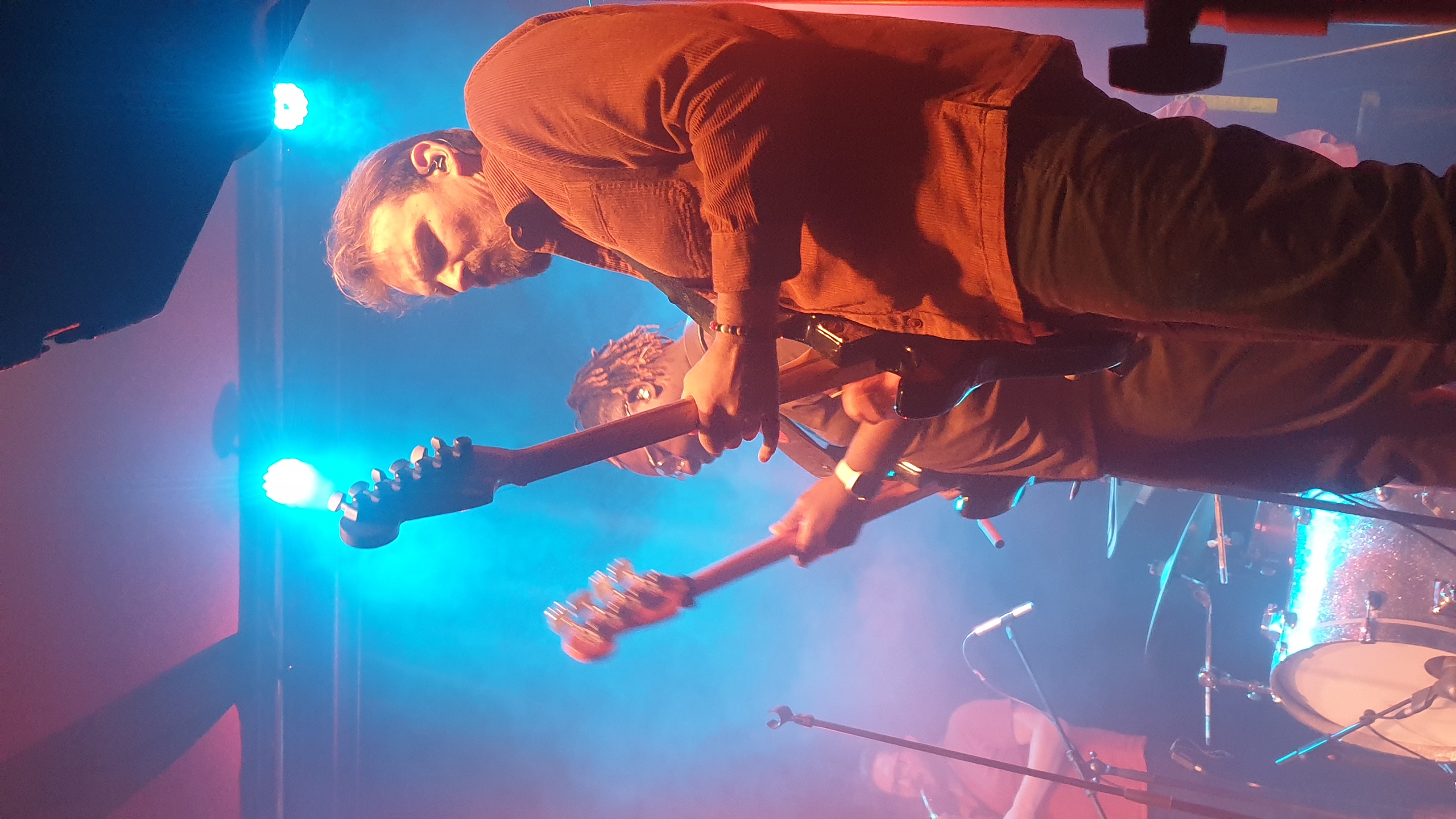 Deux musiciens du Sounds Jazz Club jouent de la guitare électrique sur scène sous des projecteurs bleus et roses intenses, avec batterie visible en arrière-plan et ambiance de concert live.