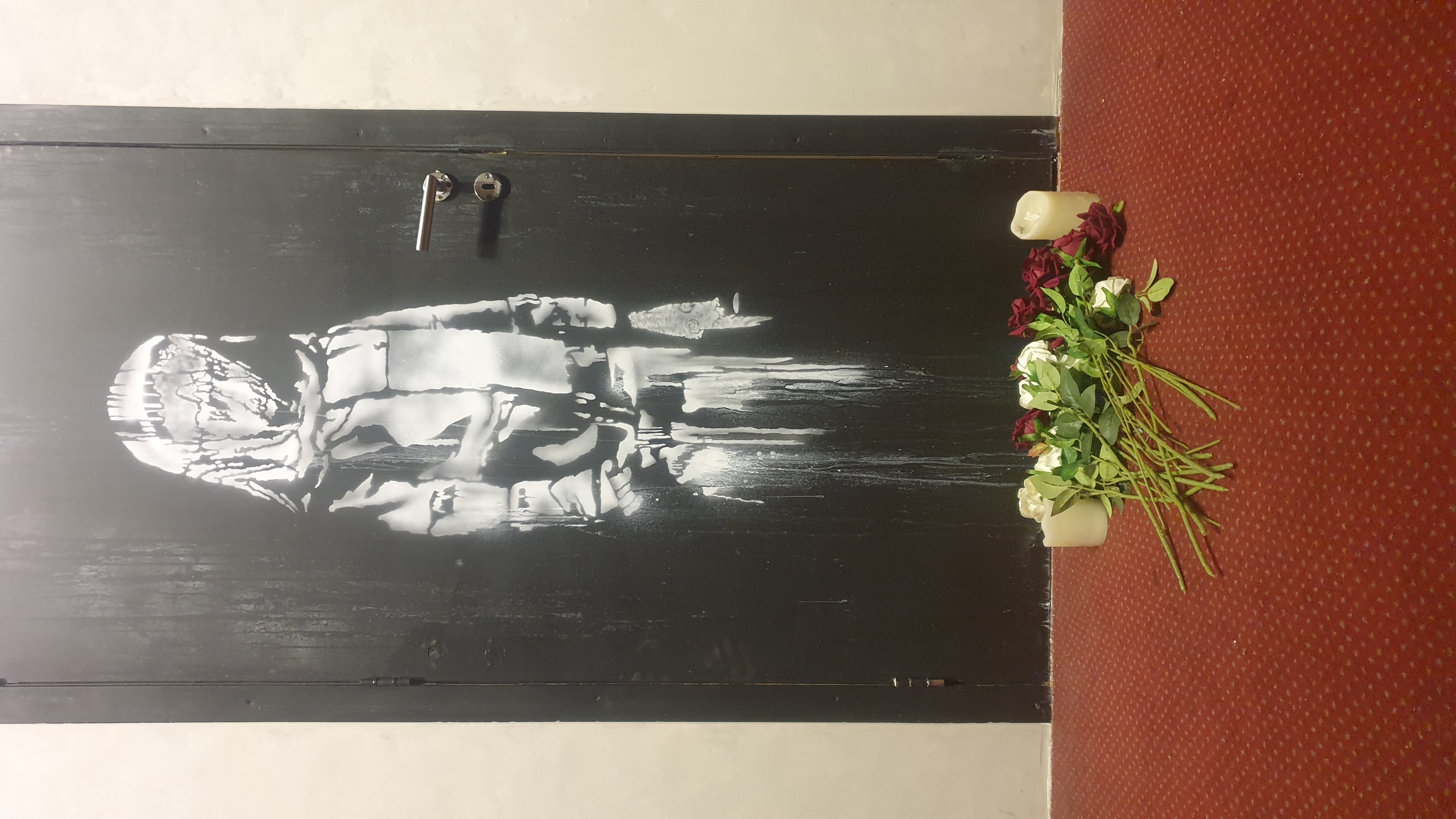 Lors de la visite du Musée Banksy en 2023. Photographie documentaire montrant une œuvre murale stencil en noir et blanc représentant une figure humaine stylisée sur un mur sombre, avec des fleurs rouges et blanches et des bougies déposées au sol en hommage, prise par Jérôme Selosse à Bruxelles.
