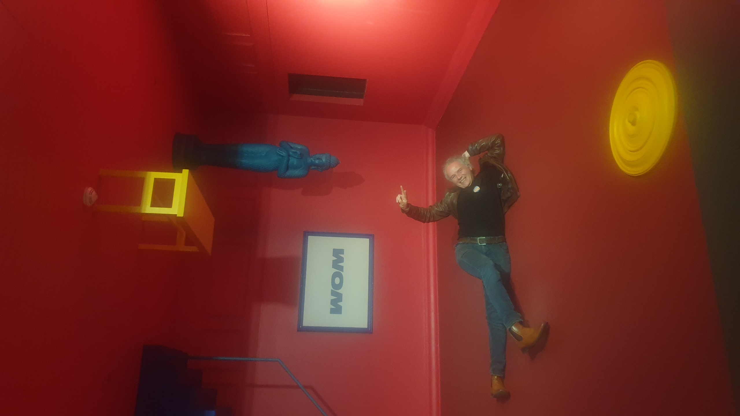 Au Wom, Jérôme Selosse allongé au sol dans une galerie aux murs rouge vif, pointant vers des éléments luminescents jaunes et des objets suspendus, créant une expérience immersive de l'artiste plasticien numérique Jérôme Selosse.