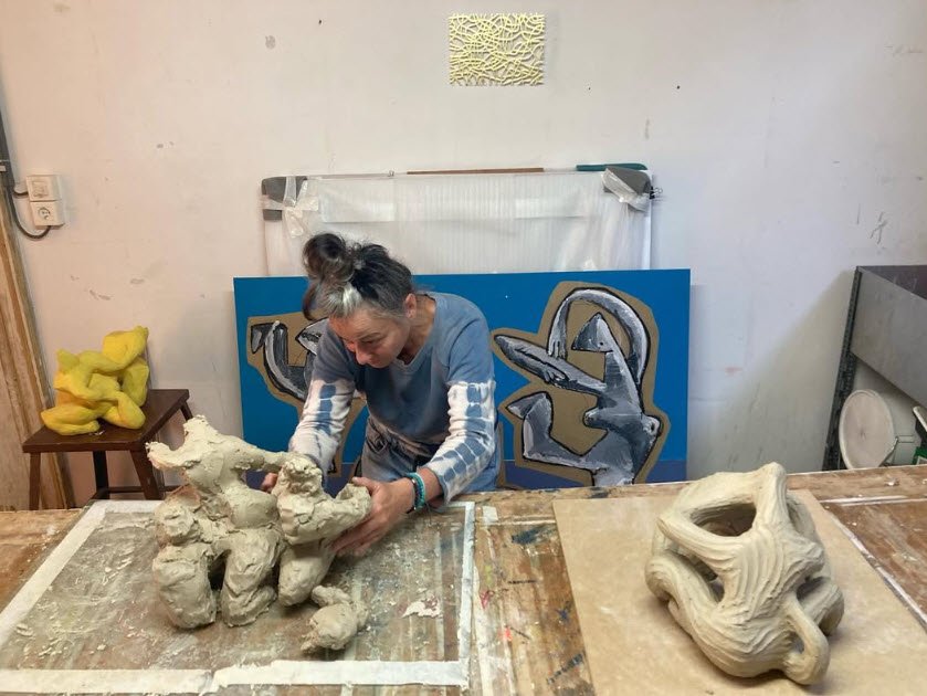 L'artiste sculptrice Gundi Falk travaille la terre dans un atelier bruxellois, les mains modelant une forme organique sur une table, avec des murs bleus en arrière-plan et des outils d'atelier visibles.

La sculptrice Gundi Falk au travail dans son atelier bruxellois, modelage de terre.