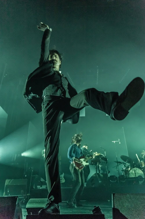 Feu!Chatterton en costume foncé saute en l'air, une jambe levée, un poing fermé dirigé vers le ciel, sous des lumières vertes de concert à L'ancienne Belgique 2019. Cliché de Lara Herbinia