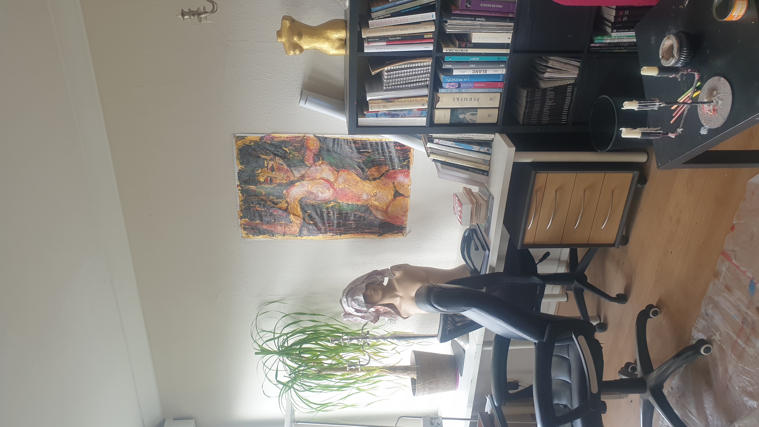Vue d'un atelier d'artiste bruxellois présentant un bureau blanc avec chaise noire, une bibliothèque remplie de livres, une toile expressionniste représentant une figure féminine aux tons chauds accrochée au mur, un buste blanc, une plante verte et des accessoires artistiques sur une table noire.