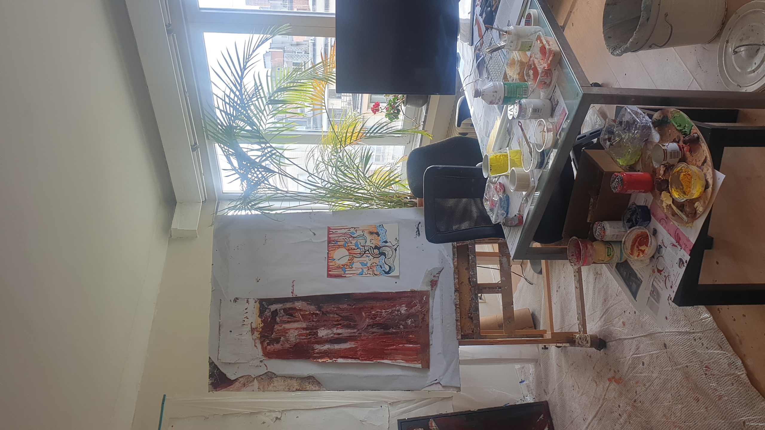 L'atelier bruxellois de SELOSSE ART, artiste et performeur, montre un espace de travail lumineux avec des toiles abstraites rouges sur chevalet, des palettes de peinture colorées, des tables de travail couvertes de matériel artistique, une plante verte en pot près de la fenêtre panoramique offrant une vue urbaine.