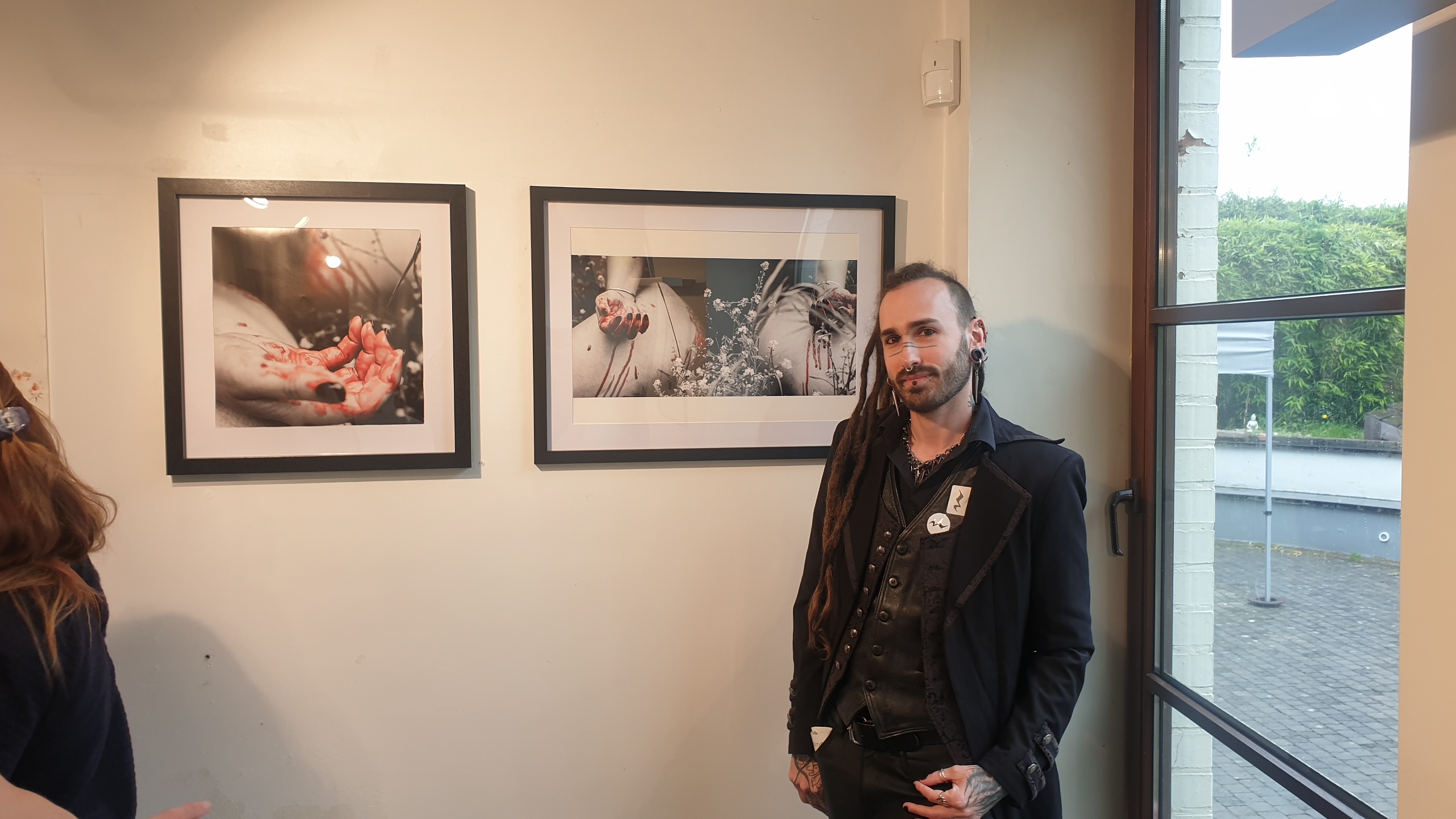 SELOSSE ART, artiste performeur bruxellois VAIN, pose devant deux photographies encadrées en galerie montrant des mains en interaction avec des éléments naturels et floraux.