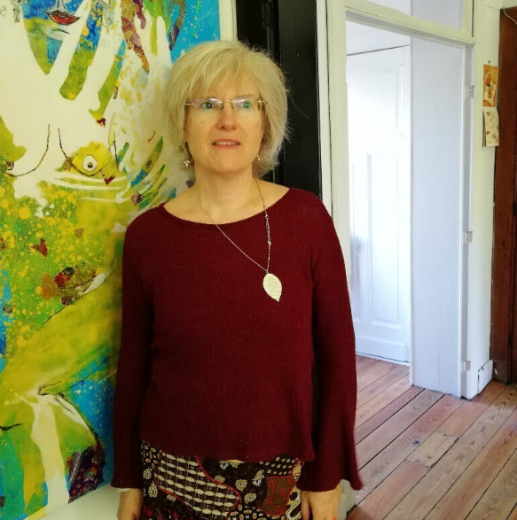 Christel Totté se tient devant une grande toile aux motifs abstraits et colorés (bleu, jaune, vert) dans un atelier avec parquet apparent et portes blanches en arrière-plan. 

Portrait de Christel Totté dans son atelier bruxellois