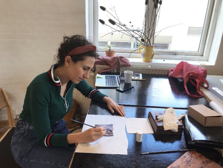 Dareen Abbas, plasticienne bruxelloise, travaille à son bureau d'atelier en dessinant sur une feuille blanche, entourée de matériaux de création et de plantes vertes près d'une fenêtre.