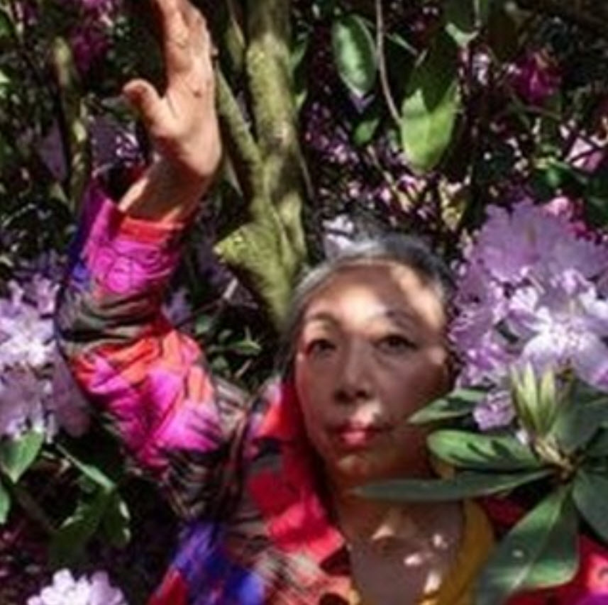 Sachiyo Honda vêtue de vêtements aux teintes vibrantes (rose, rouge, orange) est immergée parmi de grandes fleurs violettes et blanches en pleine floraison, levant le bras dans un geste de joie ou de connexion avec la nature. Portrait de l'artiste - 2024