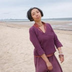 Sachiyo Honda vêtue d'un cardigan prune se tient debout sur une plage de sable, face à la mer, dans une pose contemplative.