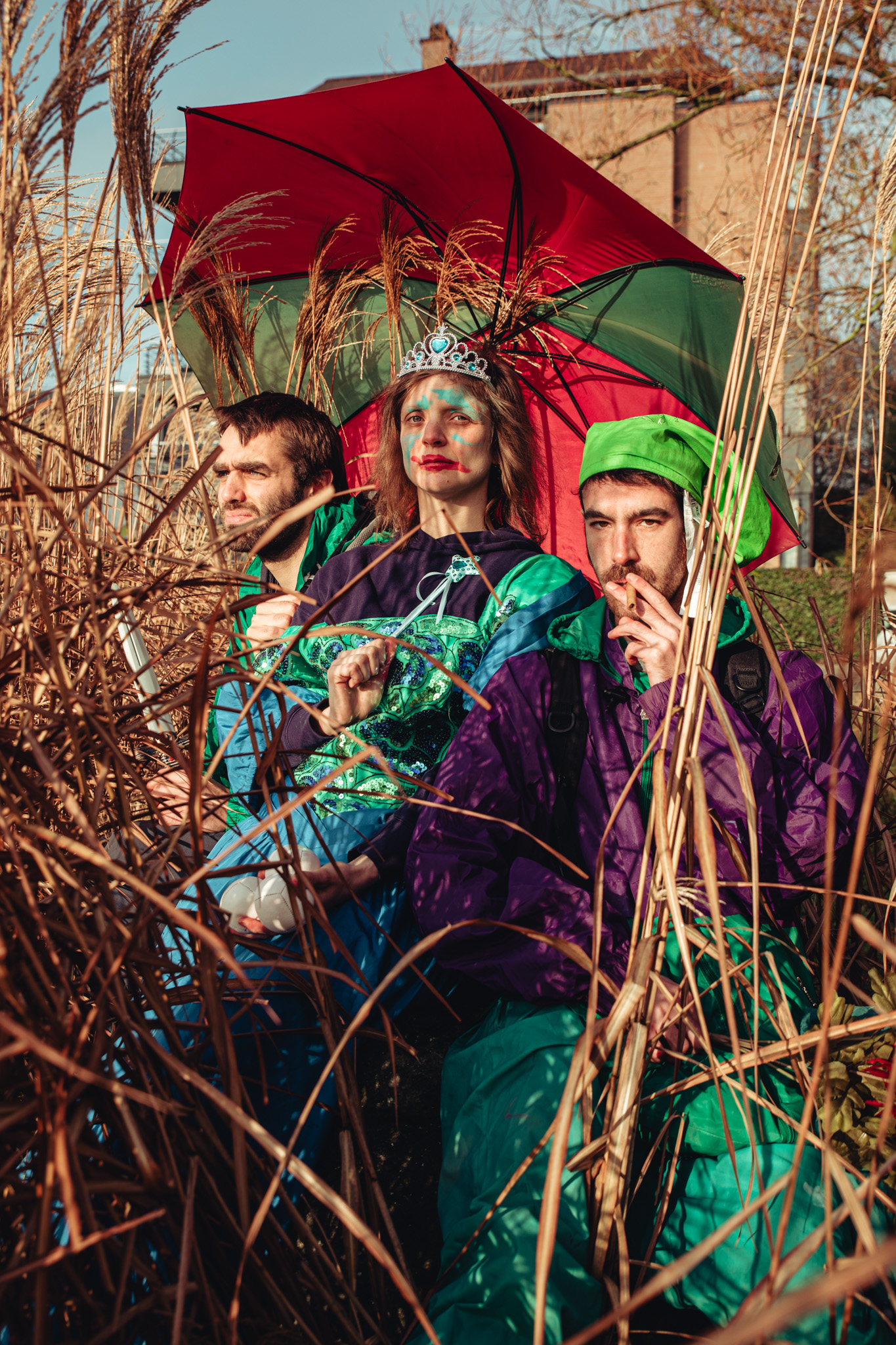 Cette photographie promotionnelle, signée Michiel Devijver, présente un portrait original et teinté d'absurde des trois comédiens de la Compagnie Anoraks pour leur création intitulée Drache Nationale.
Une parodie de conte de fées sous la pluie
Les trois artistes posent blottis les uns contre les autres au milieu de hautes herbes sèches et dorées, évoquant un abri de fortune en pleine nature. Au-dessus d'eux, un grand parapluie bicolore rouge et vert est déployé, symbolisant avec humour la drache, cette météo pluvieuse si caractéristique de la Belgique, même sous un soleil radieux.
Au centre, la comédienne incarne une sorte de princesse moderne et désabusée. Elle porte une tiare étincelante sur la tête et tient une baguette magique, mais son visage est marqué par un maquillage volontairement grossier aux nuances bleues et rouges.
À sa gauche, un comédien au regard fixe arbore un bonnet de nuit vert pomme assorti à son pantalon, tandis qu'il porte une veste de pluie violette et tient une cigarette à la bouche. À sa droite, le troisième membre du groupe, vêtu d'un manteau vert, scrute l'horizon d'un air soucieux.
L'art de l'absurde au programme de la rentrée culturelle
Ce portrait résume l'univers singulier de la Compagnie Anoraks. Entre déguisements hétéroclites, attitudes théâtrales et poésie du quotidien, le trio s'amuse à détourner les codes de la météo et des récits d'aventures traditionnels.
Ce spectacle, proposé lors de la Joyeuse Rentrée d'Ixelles au Petit Théâtre Mercelis, promet d'apporter une bonne dose d'autodérision et de rire aux spectateurs venus célébrer le lancement de la saison artistique 2026.