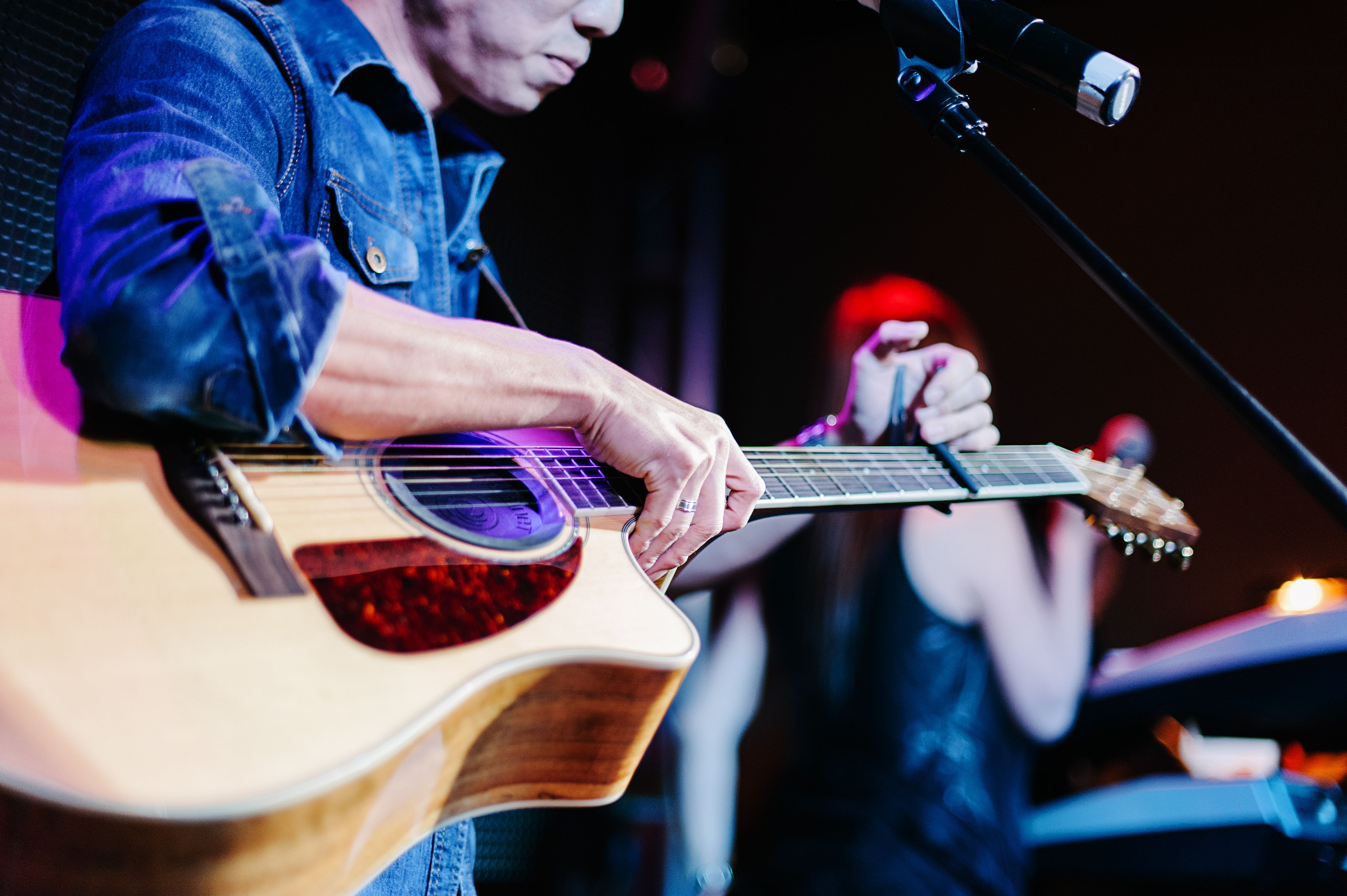 Cette image saisit l'intimité d'une performance musicale sur scène, illustrant la diversité des concerts proposés lors de la rentrée culturelle.

La concentration et la précision du musicien
Au premier plan, un guitariste vêtu d'une chemise en jean bleu ajusté se concentre sur sa guitare acoustique. Ses mains, capturées de près, sont en train de positionner un capodastre sur le manche de l'instrument pour en modifier la tonalité. La guitare folk présente une table en bois clair et une plaque de protection ambrée, typique des instruments de scène traditionnels.

Un microphone noir fixé sur son pied s'avance dans le coin supérieur droit, prêt à capter chaque note et chaque nuance de l'instrument.
Une atmosphère de scène chaleureuse
L'arrière-plan, volontairement flou grâce à une faible profondeur de champ, révèle une seconde artiste sous des éclairages de scène chaleureux aux teintes violettes et rouges. Sa silhouette suggère un duo ou un groupe en pleine synergie créative, renforçant la sensation de complicité propre aux spectacles en direct.

Cette esthétique chaleureuse et centrée sur l'instant présent évoque l'expérience des concerts intimistes programmés pour la Joyeuse Rentrée. C'est notamment le cas des sessions acoustiques et des découvertes de musiques du monde proposées par l'association Muziekpublique sur la Place Fernand Cocq. 

Ce gros plan rappelle l'échange authentique entre les artistes locaux et leur public, réunis autour de la passion de la musique vivante.