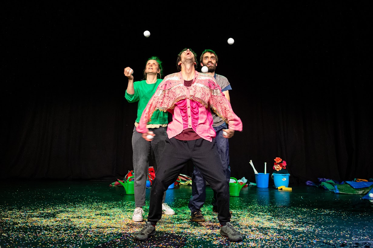 Cette photographie, créditée à Thomas Andrien, saisit une scène dynamique et pleine de malice issue du spectacle Drache Nationale, présenté par la Compagnie Anoraks lors de la Joyeuse Rentrée à Ixelles.

Une performance physique sous le signe de l'humour
Sur une scène au fond noir, trois comédiens partagent l'espace dans une chorégraphie clownesque. Au centre, un comédien vêtu d'une chemise rose originale ornée de dentelle blanche jette sa tête en arrière dans une attitude de concentration intense. Autour de lui, plusieurs balles blanches de jonglage semblent suspendues dans les airs.

À sa gauche, une comédienne aux cheveux teintés en vert, habillée d'un pull vert vif, participe activement à cette prouesse physique en suivant elle aussi des yeux le mouvement des balles. Derrière eux, un troisième artiste observe l'action avec une expression captivée, prêt à intervenir.
Un univers scénique coloré et décalé
Le sol de la scène est recouvert d'une multitude de confettis multicolores, apportant une dimension festive et désordonnée au décor. À l'arrière, divers objets ordinaires sont disposés au sol, comme des arrosoirs en plastique vert, des seaux bleus et des fleurs artificielles. Ces accessoires font directement écho au thème de la drache, ce terme typiquement belge utilisé pour désigner une forte averse de pluie, que la compagnie transforme ici en un terrain de jeu poétique.

Ce spectacle, programmé à 15h00 au Petit Théâtre Mercelis, illustre la volonté d'Ixelles de proposer des formes de spectacle vivant accessibles, à la fois drôles et touchantes, pour inaugurer la rentrée culturelle 2026.