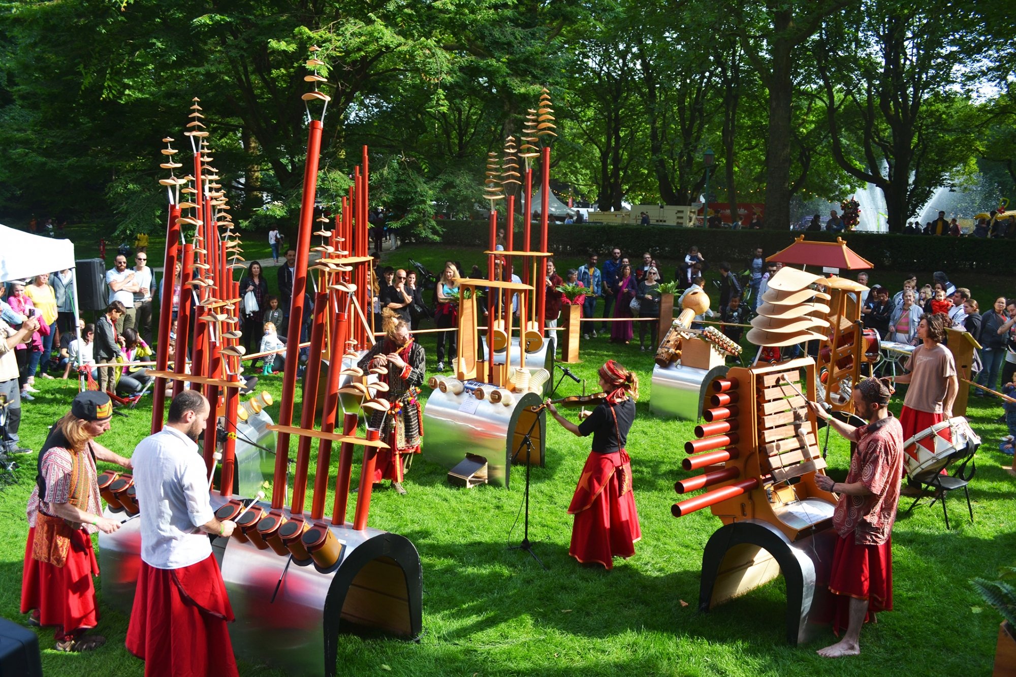 Cette image présente l'installation sonore Fujinn, créée par le collectif Amadeo, en pleine représentation au cœur d'un parc verdoyant. Il s'agit d'un ensemble de sculptures musicales géantes et insolites, conçues pour être jouées à la fois par des artistes et par le public de passage.

Une architecture sonore unique et colorée
Au milieu d'une pelouse bien verte, plusieurs structures hybrides captent immédiatement le regard. Elles se composent de socles en métal cintré brillant sur lesquels reposent de longs tubes verticaux peints en rouge vif. Ces tubes sont surmontés de fines pièces en bois empilées, rappelant des structures végétales ou des éléments de pagodes orientales.

Chaque sculpture intègre des instruments différents. Sur l'une d'elles à droite, on aperçoit un xylophone en bois intégré, tandis que d'autres disposent de petits tambours, de cymbales, de cloches ou de claviers.

Une performance vivante et participative
Plusieurs artistes vêtus de tenues aux tons rouges et de style artisanal animent l'espace en jouant de leurs instruments respectifs. On distingue notamment une violoniste au centre, un flûtiste, ainsi que des percussionnistes qui frappent le rythme directement sur les modules intégrés aux structures métalliques.

À l'arrière-plan, un public familial s'est rassemblé en demi-cercle sous l'ombre de grands arbres pour observer le spectacle. Une fontaine d'eau visible au loin complète ce tableau bucolique et détendu.

Dans le cadre de la Joyeuse Rentrée culturelle à Ixelles, cette œuvre interactive invite les petits comme les grands à s'approprier ces sculptures poétiques pour improviser, manipuler les matières et créer ensemble une symphonie spontanée.