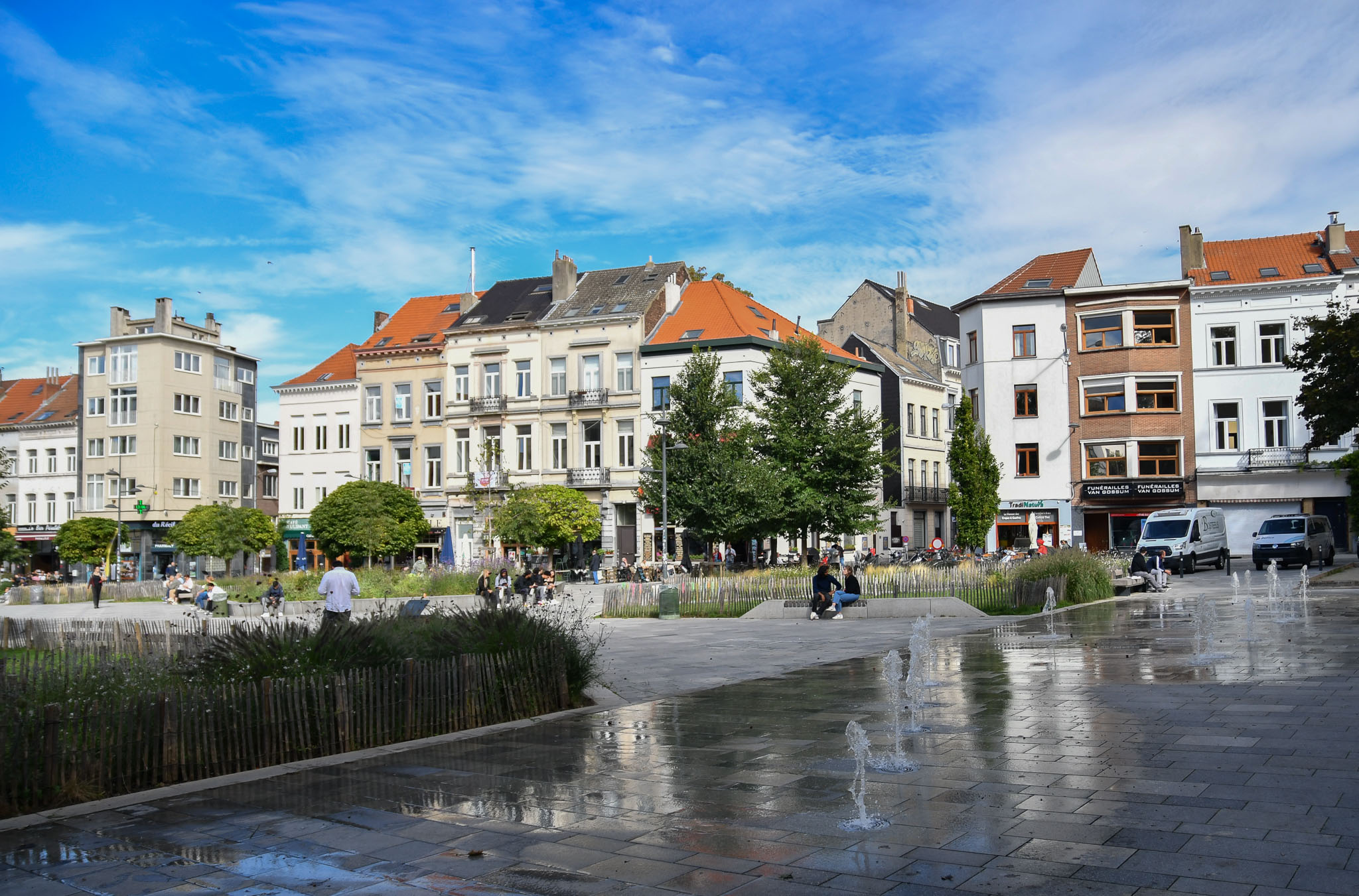 Un décor ouvert et convivial au cœur d'Ixelles
La photographie présente la Place Fernand Cocq sous un ciel d'un bleu lumineux parsemé de légers nuages blancs. Cette esplanade moderne et piétonne se révèle être le cadre idéal pour accueillir un grand festival en plein air.

Au premier plan, le sol pavé et humide reflète la lumière du jour ainsi que les jets d'eau verticaux qui s'élancent de manière ludique. Cet espace dégagé et dynamique offre une belle surface pour les performances de rue, notamment pour les déambulations sonores de la fanfare BRUiTAL ou l'installation de la sculpture sonore interactive Fujinn.

Au centre de la place, des îlots de verdure entourés de ganivelles, ces petites barrières en bois rustiques, apportent une touche naturelle et chaleureuse. Des bancs et des murets en pierre y sont aménagés, invitant les spectateurs à s'installer confortablement pour apprécier les concerts, comme celui du Brussels Film Orchestra, ou pour simplement profiter de l'ambiance festive en famille entre deux animations.

En arrière-plan, les façades typiques de l'architecture bruxelloise s'alignent avec leurs différents styles et couleurs. Les commerces au rez-de-chaussée soulignent le caractère vivant et de proximité de ce quartier ixellois. C'est précisément cette atmosphère de village urbain, à la fois accessible et accueillante, qui caractérise l'esprit de la Joyeuse Rentrée culturelle de 2026.