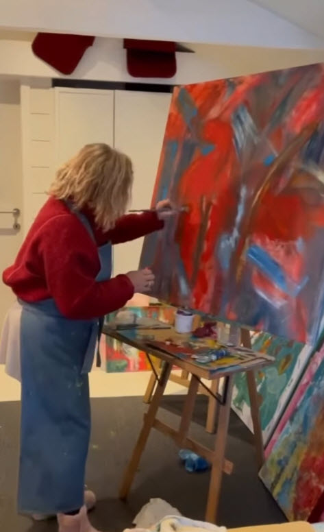 Sacha Valsamides peint sur une grande toile aux teintes rouges et bleues, debout devant son chevalet dans un atelier bruxellois lumineux, palette et pinceaux à portée de main.