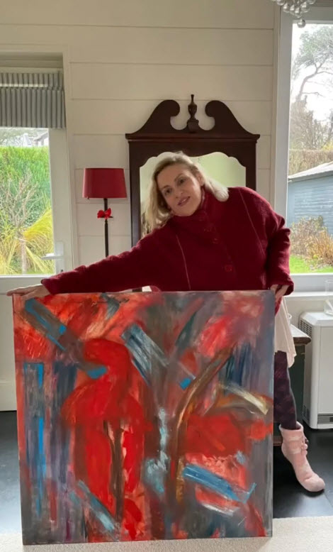 Sacha Valamides tient une peinture abstraite aux tonalités rouges, bleues et grises devant elle, dans un intérieur lumineux bruxellois avec mobilier ancien en arrière-plan.