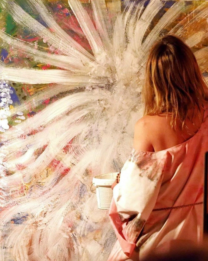 Olena Rabitcheva en train de peindre dans son atelier une peinture abstraite aux coups de pinceau blanc et rose rayonnants. Témoignage de l'artiste: C'est par la mort, la douleur et la souffrance que j'apprenais à aimer, apprécier et célébrer la vie. Et après être passée de l'autre côté à plusieurs reprises et avoir reçu la chance de continuer la vie, juste pour peut-être devenir sa plus grande admiratrice, la gardienne du patrimoine du vivant et du beau et unique.

Face à la mort, tu as souvent le regret de n'avoir pas osé vivre pleinement, sans peur, pour sublimer le créateur divin, son code unique qu'il a mis en toi. De n'avoir pas assez su la valoriser et crier très haut partout : regardez cette graine magique que Dieu a mise en moi, qui fait tellement de choses incroyables et jamais encore existé dans ce monde. Et inspirer les autres à cultiver leur graine magique. De savoir fermer les portes très vite à la tentation qui veut te dévier de ton chemin et prendre ton temps, ton énergie et tes ressources juste pour te ralentir et t'empêcher d'être cette étoile telle que Dieu t'a créée.
Text Olena R gallerie