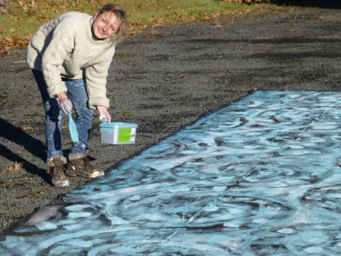 Photographie de Olena Rabitcheva, vêtue d'un pull beige et de jeans, en pleine action elle peint une grande toile bleue posée au sol avec un pinceau, tenant un pot de peinture vert à la main.