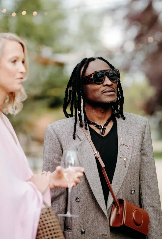 Un moment de détente dans une soirée privée. Chibida Dumbu portant des lunettes noires, un blazer gris et un sac marron, regarde vers le haut tandis qu'une femme en rose se tient à ses côtés. Bokeh de lumières dorées en arrière-plan.

Chibida Dumbu, musicien de Bruxelles. Un moment de détente dans une garden party.

Moment extrait de son insta: https://www.instagram.com/p/DZM3f4jAkWs/?img_index=2