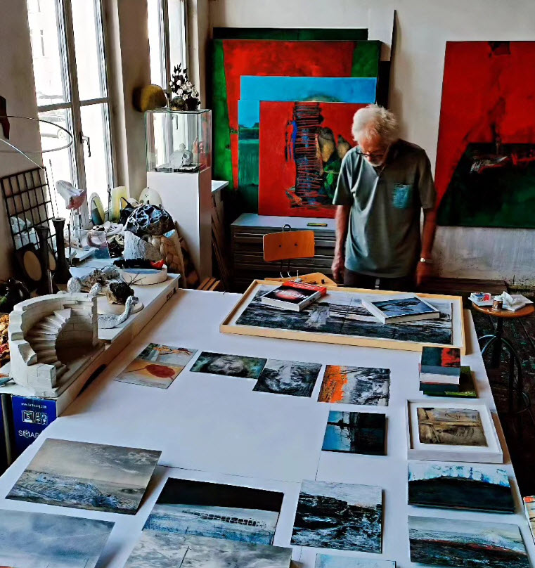 Christian Rolet l'artiste peintre, examine des tirages photographiques disposés sur une table dans son atelier, entouré de grandes toiles colorées aux teintes de rouge, bleu et vert.