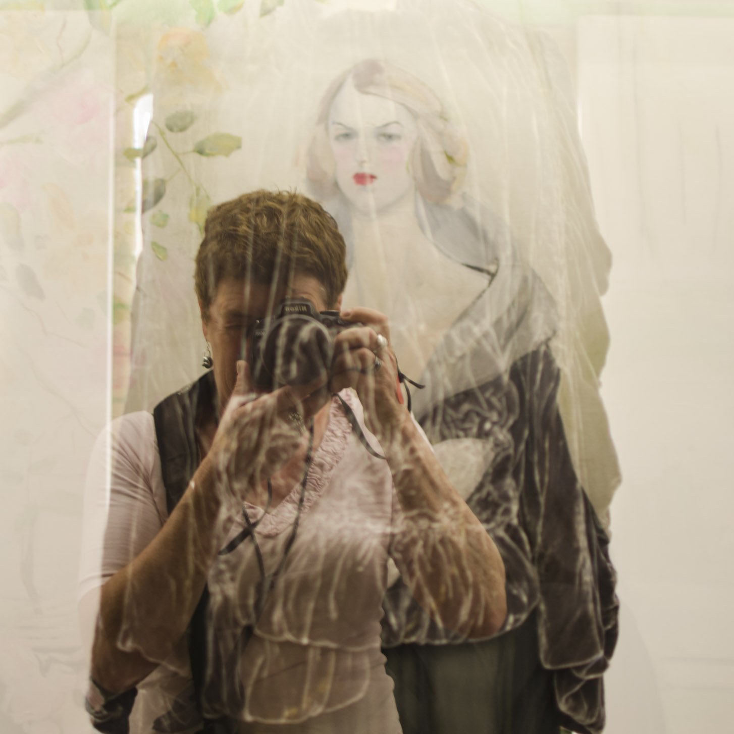 Une personne tenant un appareil photo devant une grande illustration murale représentant une femme aux cheveux longs et aux lèvres rouges, dans une galerie aux murs clairs.