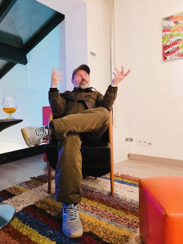 Damien Paul Gal (DPG) assis sur une chaise en bois, les bras levés expressifs, dans un espace d'exposition avec des œuvres colorées et un tapis multicolore au sol.

Reportage photo de Damien-Paul Gal à l'exposition collective de peinture hôte gallery Bruxelles