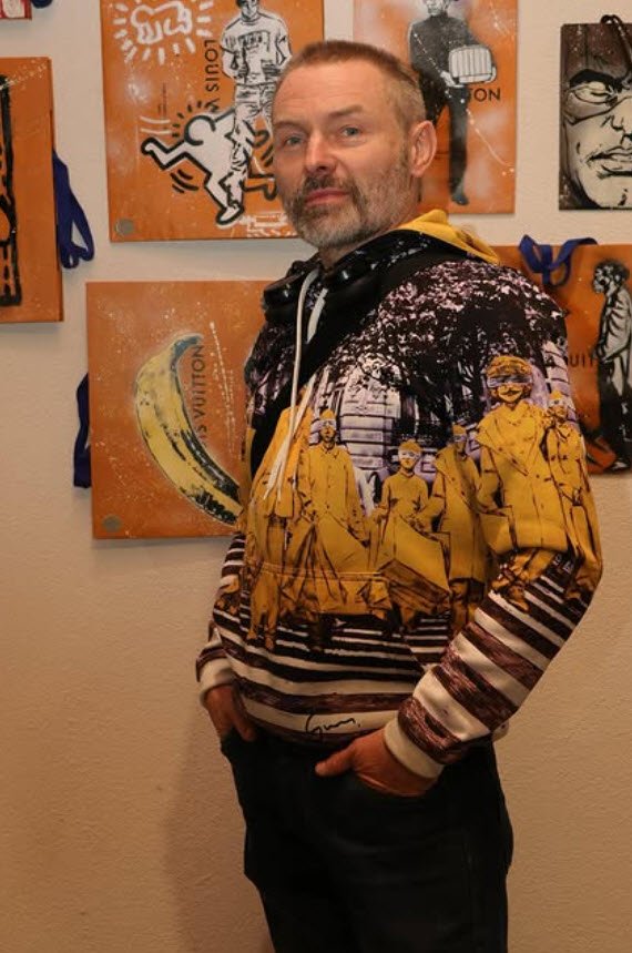 Portrait de Damien-Paul Gal, plasticien et street artist, portant un hoodie aux motifs graphiques jaunes et noirs, posant devant ses œuvres d'art affichées sur des panneaux orange en arrière-plan.

Portrait de l'artiste Damien-Paul Gal dans son univers graphique