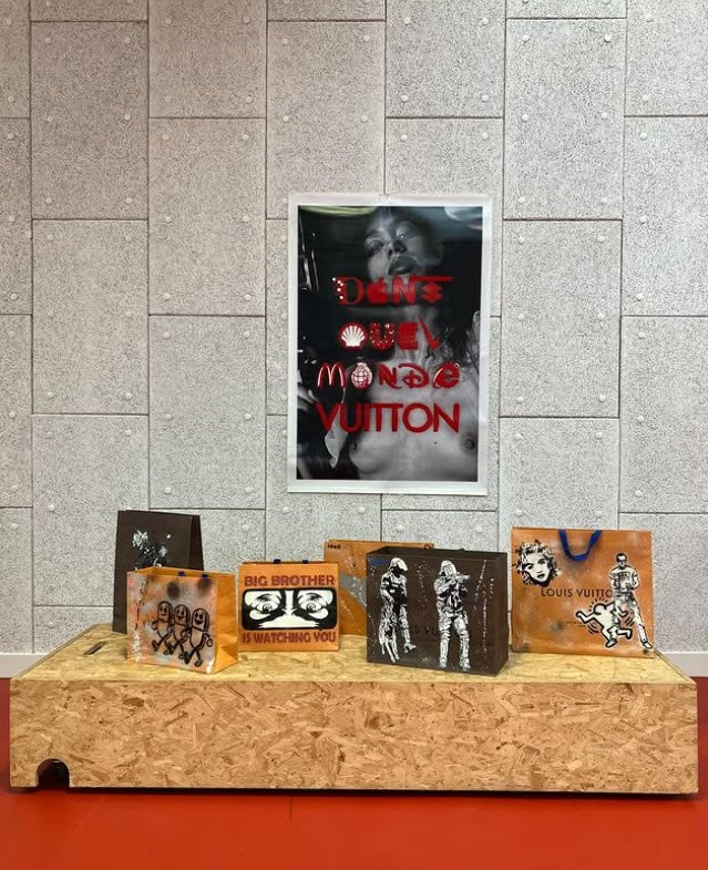 Installation de Damien-Paul Gal exposant des sacs peints en orange et noir ornés de logos détournés, disposés sur un socle en liège brut face à un mur de carrelage gris, avec une affiche neon rouge suspendue au-dessus montrant un visage superposé de logos de marques de luxe, à Bruxelles.

Reportage photo de Damien-Paul Gal : installation « Dans quel monde Vuitton » au Parcours d'artiste de Saint-Gilles 2026, Bruxelles

Extrait insta de l'artiste: https://www.instagram.com/p/DW_SSvoCE0p/