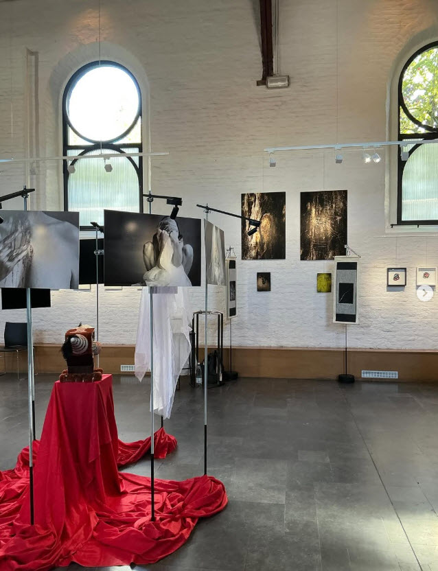 Intérieur de la chapelle de Boondael. Galerie bruxelloise aux murs blancs et hautes fenêtres arquées : installations artistiques rouge écarlate, des œuvres photographiques encadrées et des tirage noir et blanc accrochés sur les cloisons. Exposition collective Frédérique Dehoux lors du parcours d'artiste d'Ixelles 2025.