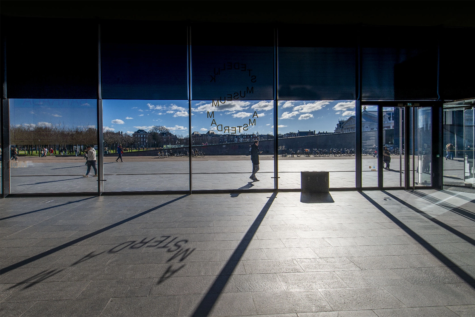 Intérieur d'un musée à Amsterdam avec larges baies vitrées offrant une vue panoramique sur une place ensoleillée, des visiteurs dispersés, et le texte inversé du mot musée visible sur la vitre.